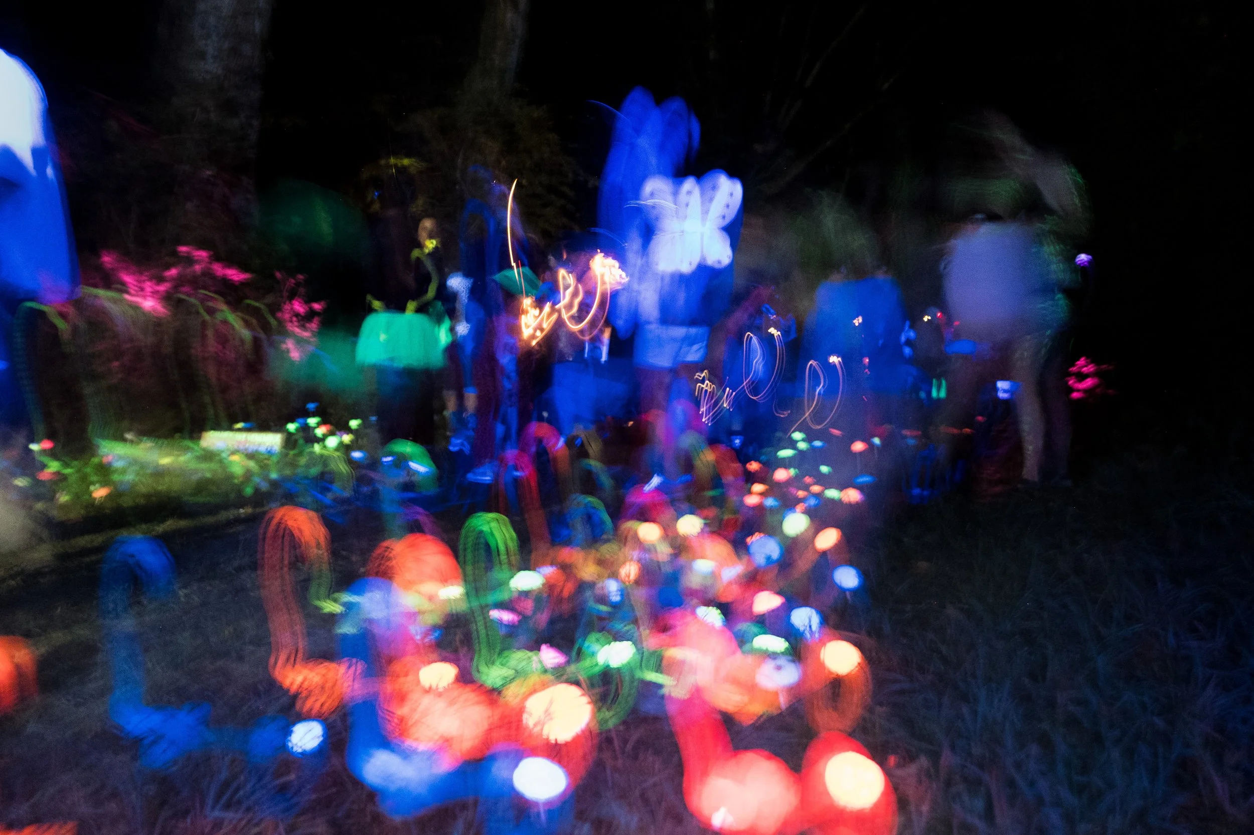 Image floue d'une fête avec des lumières colorées, probablement des néons ou des lampes LED, créant un effet d'arc-en-ciel lumineux. Des personnes sont présentes, mais leur apparence est indistincte en raison du mouvement.