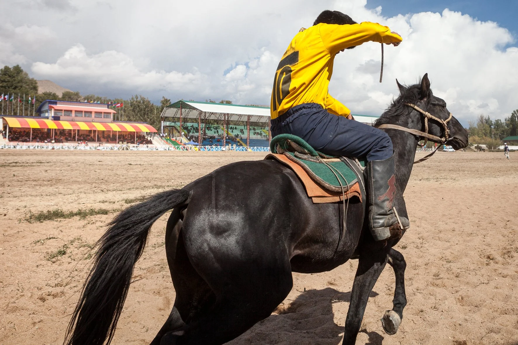 Jockey en selle sur un cheval noir lors d'une compétition de kok boru, avec tribunes et spectateurs en arrière-plan. Ciel nuageux.