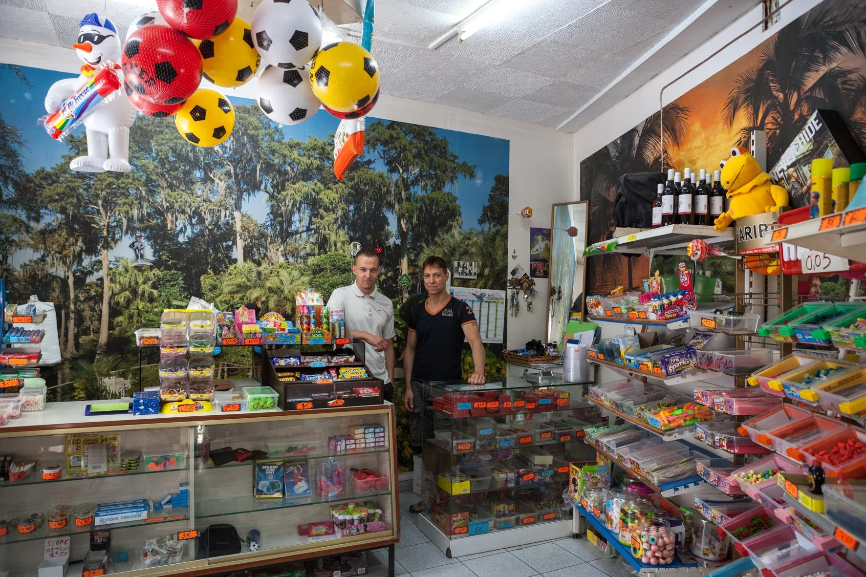 Deux personnes derrière un comptoir dans une boutique de bonbons, entourées de rayons remplis de confiseries colorées, avec un décor forestier en arrière-plan.