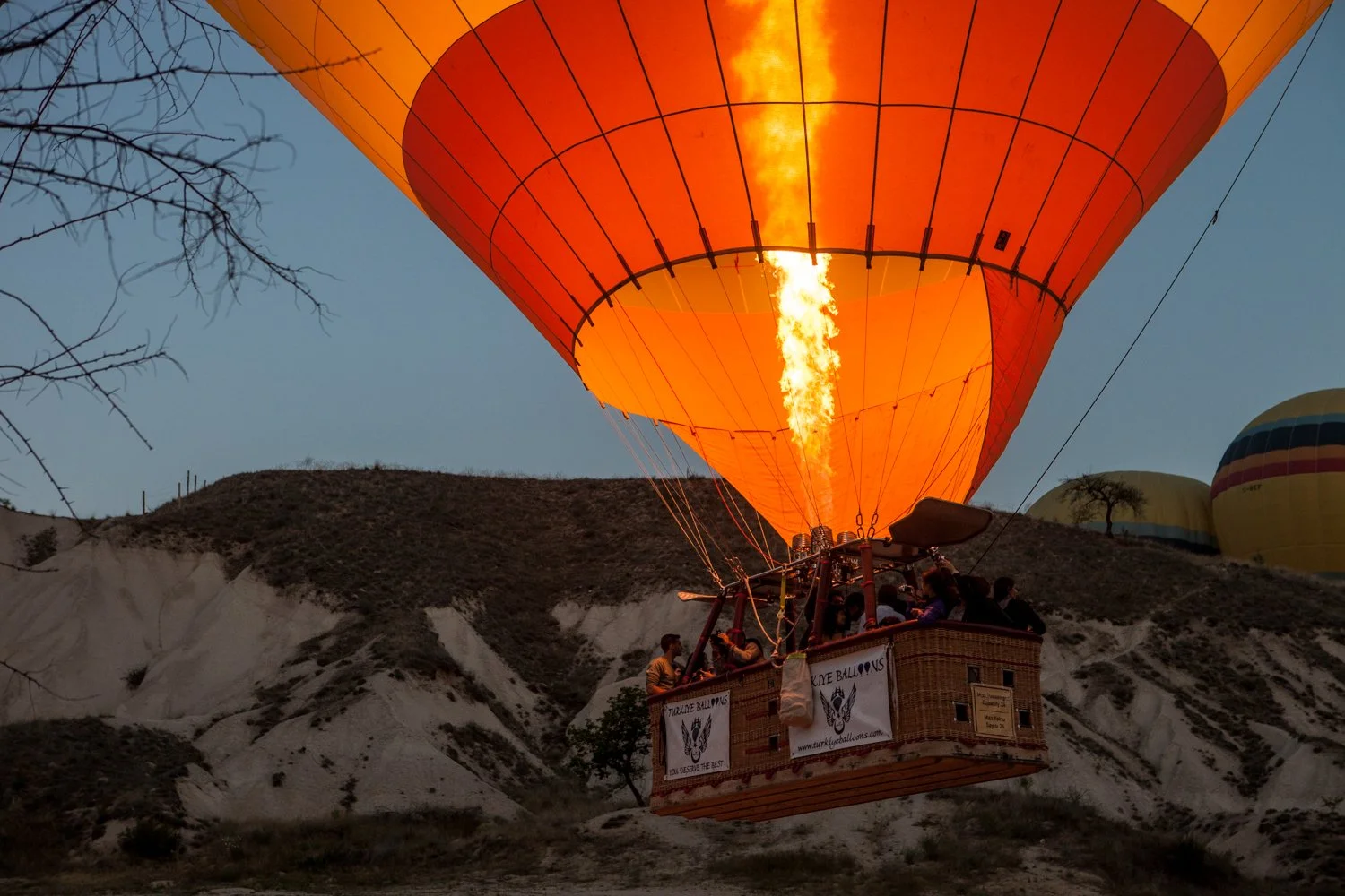 Ballon à air chaud orange en train de s'envoler avec un feu allumé en dessous, portant des passagers, dans un paysage de collines rocheuses, avec plusieurs autres ballons visibles en arrière-plan.