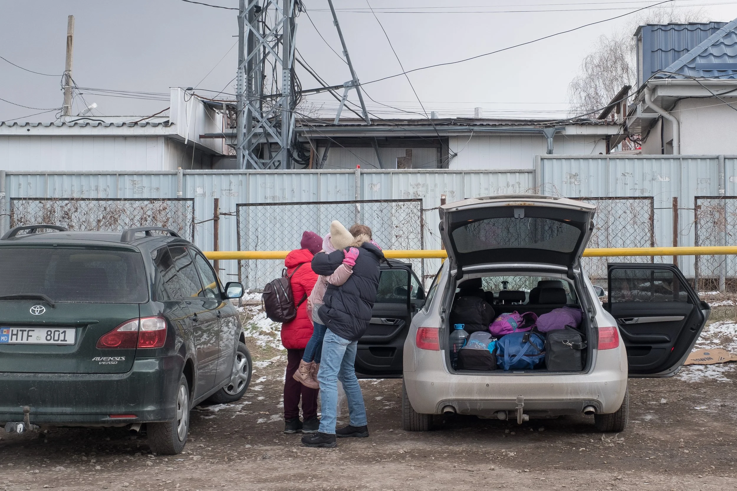 Réfugiés ukrainiens se retrouvant et s'enlaçant dans un parking avec des voitures et un mur en métal en arrière-plan.