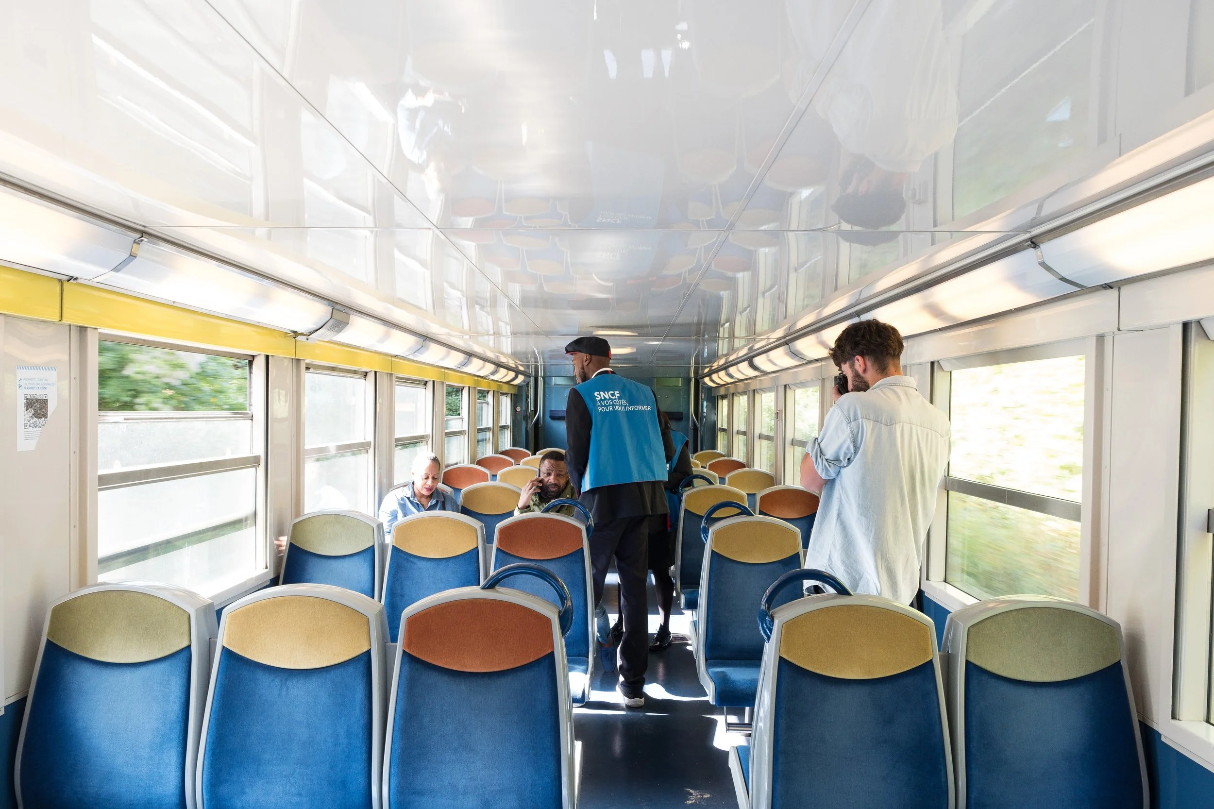 À l'intérieur d'un train, quelques passagers assis et un agent de la SNCF se tenant debout. La scène est lumineuse avec des fenêtres donnant sur un paysage vert à l'extérieur.