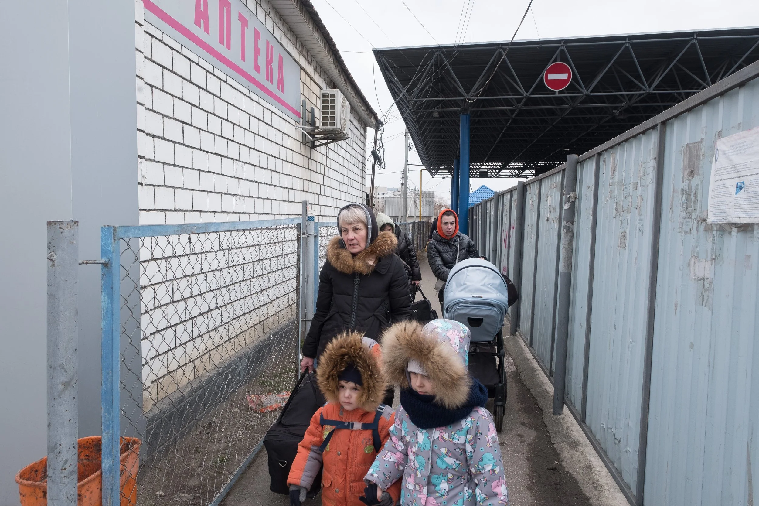 Groupe de réfugiées ukrainiennes, dont une femme et deux enfants, marchant en file dans une allée d'un poste frontière moldave délimité par des barrières métalliques sur le côté, portant des vêtements chauds avec capuches en fourrure, en plein hiver.