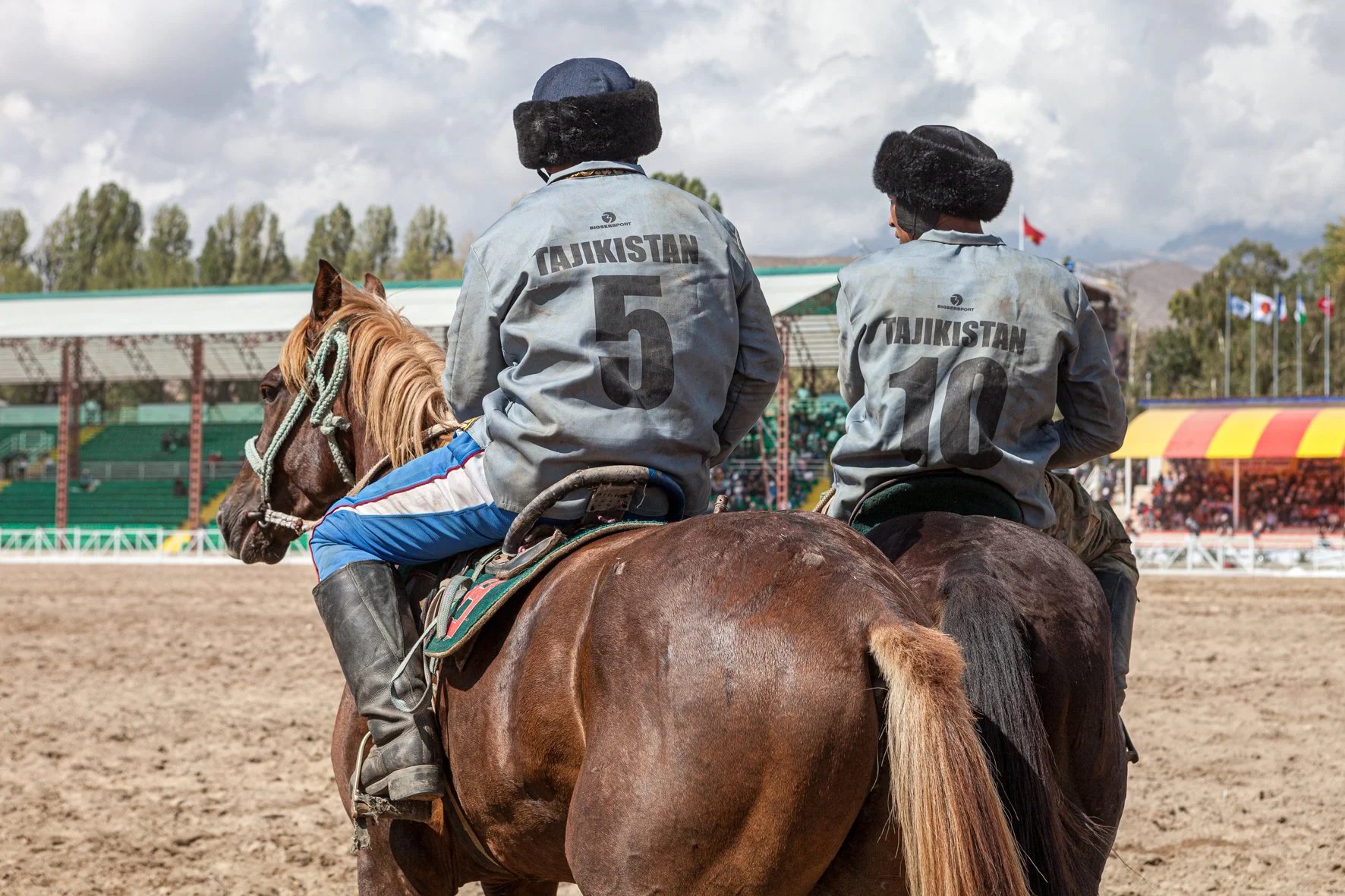 Deux joueurs de kok boru du Tajikistan, portant des vestes grises avec les numéros 5 et 10, montent leur cheval sur un terrain, avec un public et des drapeaux dans le stade en arrière-plan.