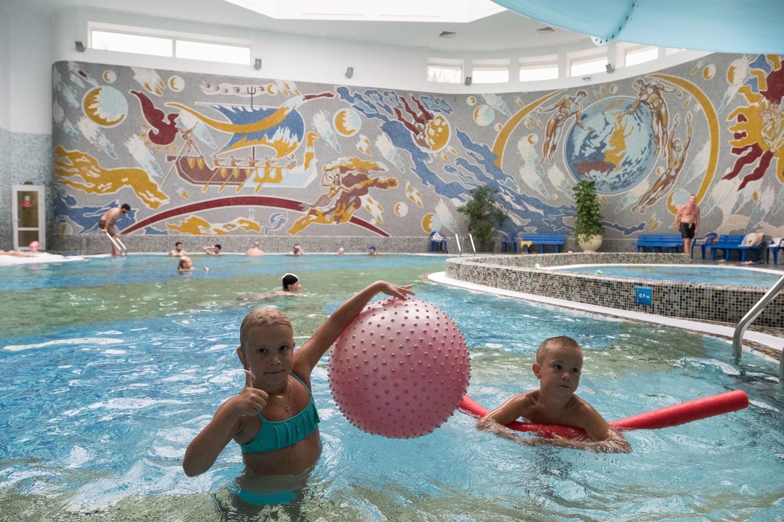 Deux enfants dans une piscine intérieure, l'un avec une boule rose et l'autre nageant avec une frite rouge. En arrière-plan, plusieurs personnes nagent et se détendent dans la piscine, une grande fresque colorée orne le mur derrière eux, à Minsk.