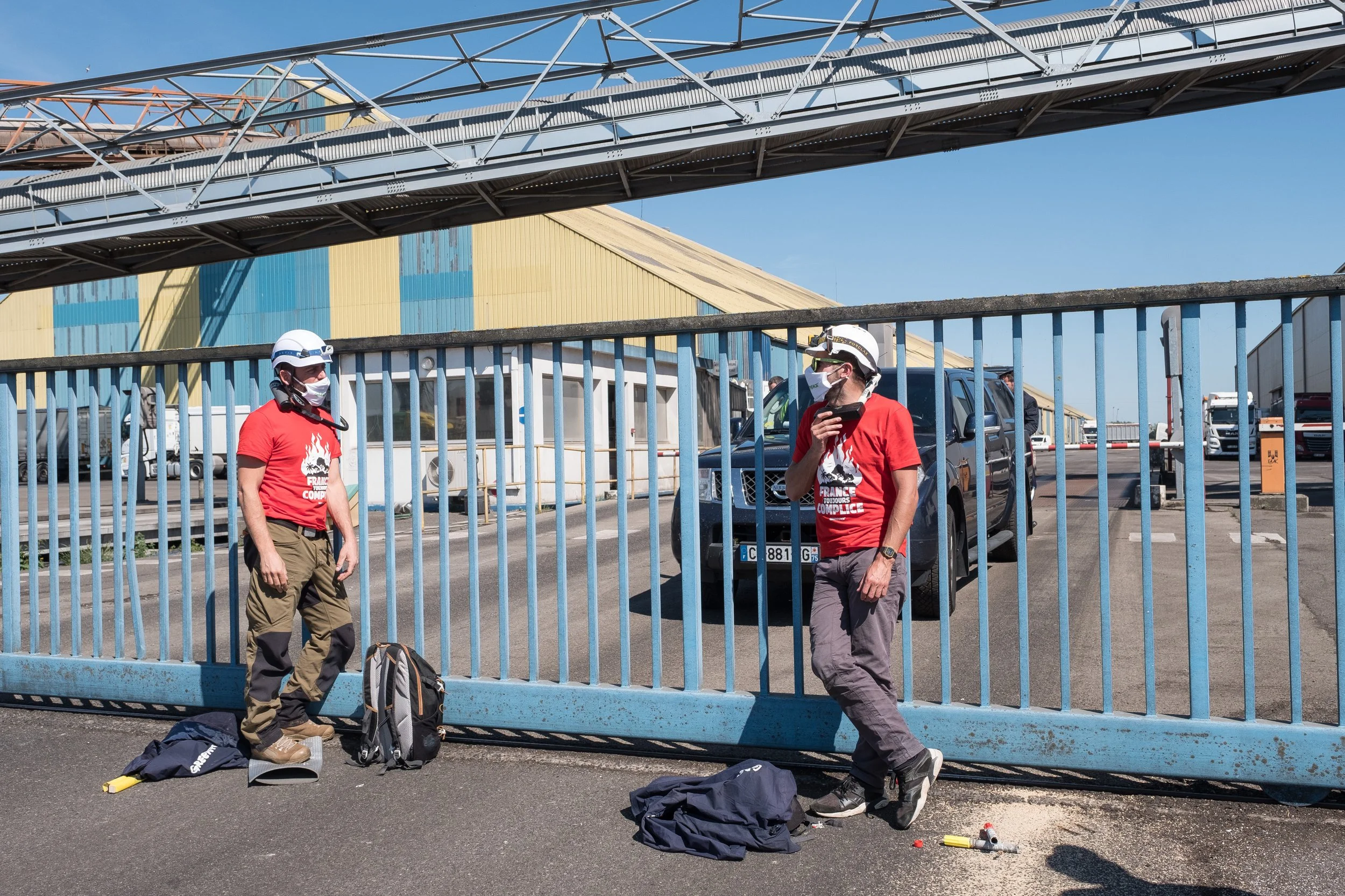 Deux activistes de Greenpeace portant des casques, masques et t-shirts rouges avec un logo de protestation, discutent à côté d'une barrière bleue à l'extérieur d'un bâtiment industriel. Il y a plusieurs voitures en arrière-plan.