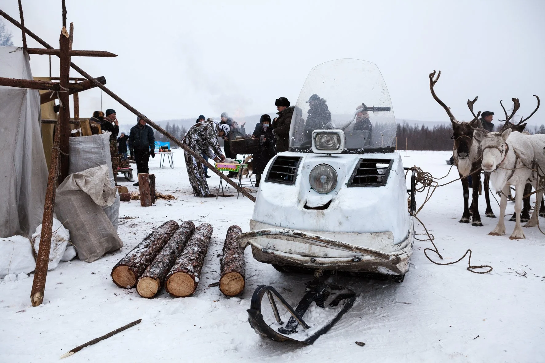 Une scène hivernale avec une moto-neige, des personnes, des chevaux, et un bâtiment en bois en arrière-plan.