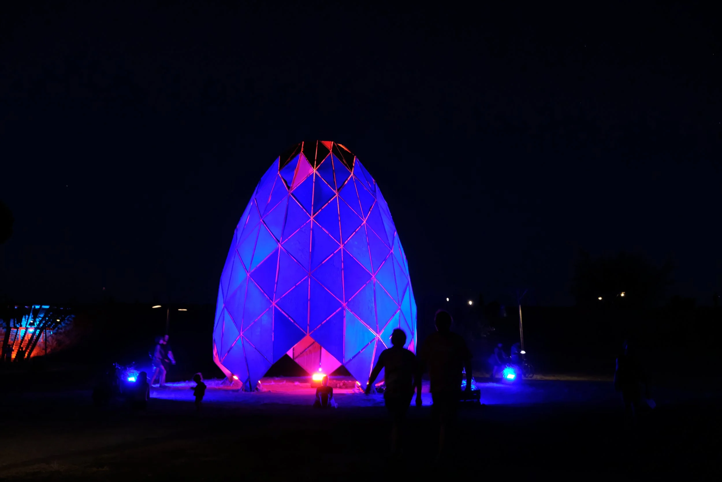 Structure en forme d'œuf illuminée de lumières bleues et rouges, visible la nuit, avec silhouettes de personnes en avant-plan au Waking Life au Portugal.