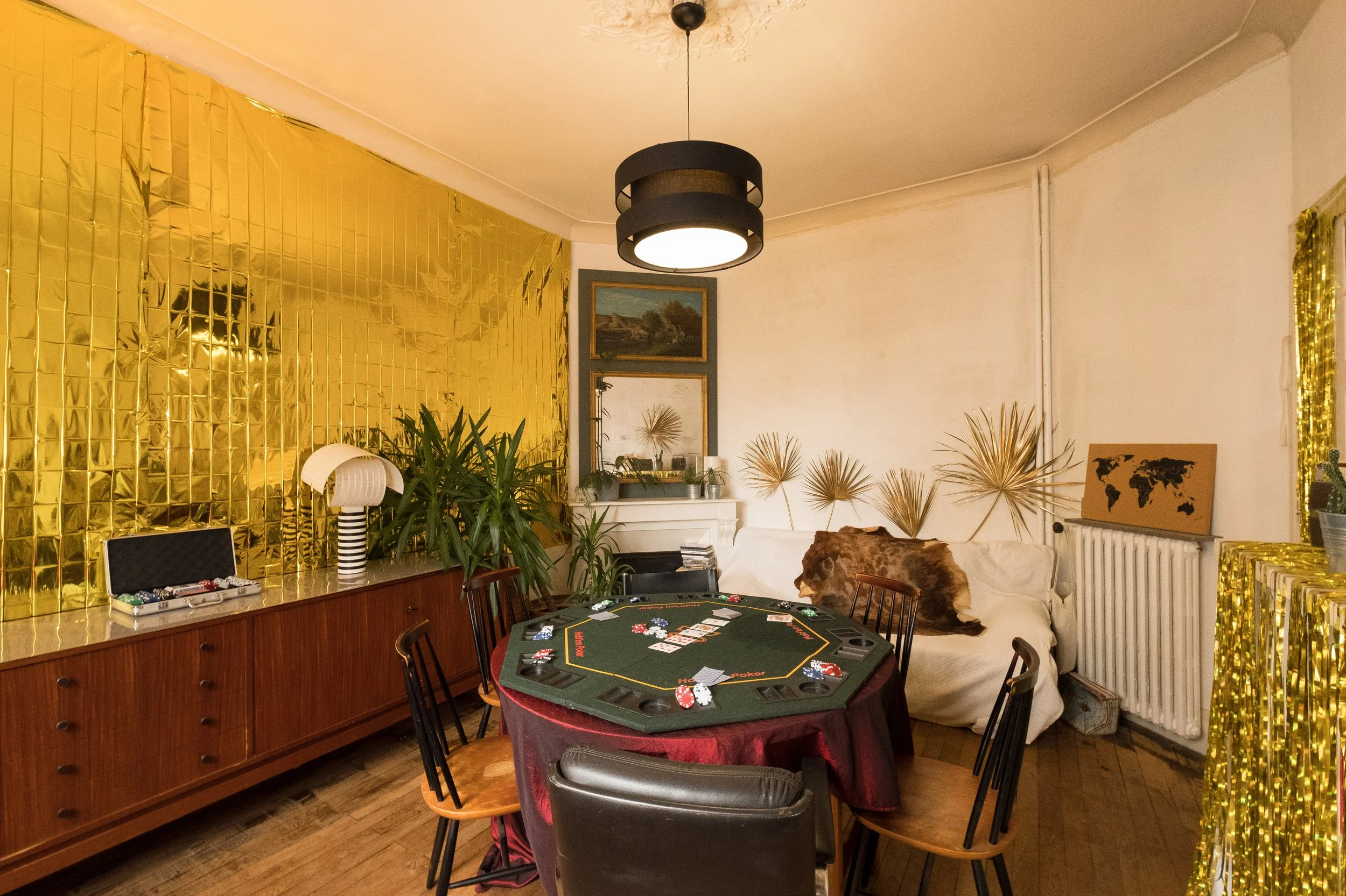 Salle de jeu avec un tapis de poker sur la table, décorations dorées, plantes vertes, un canapé blanc avec des coussins en fourrure, un buffet en bois, et deux miroirs au mur.