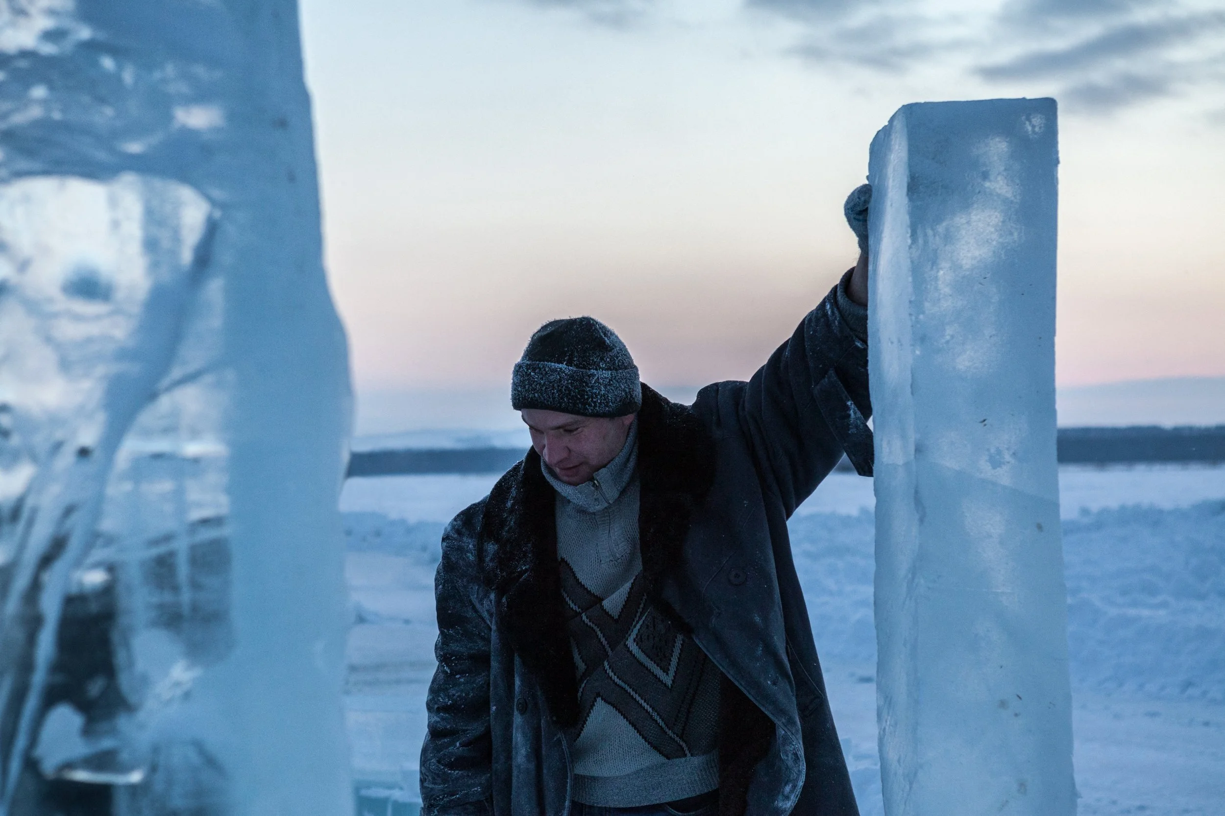 Un homme habillé chaudement en hiver, portant un bonnet, se tient dans un environnement enneigé, entre deux grandes structures de glace en Russie.