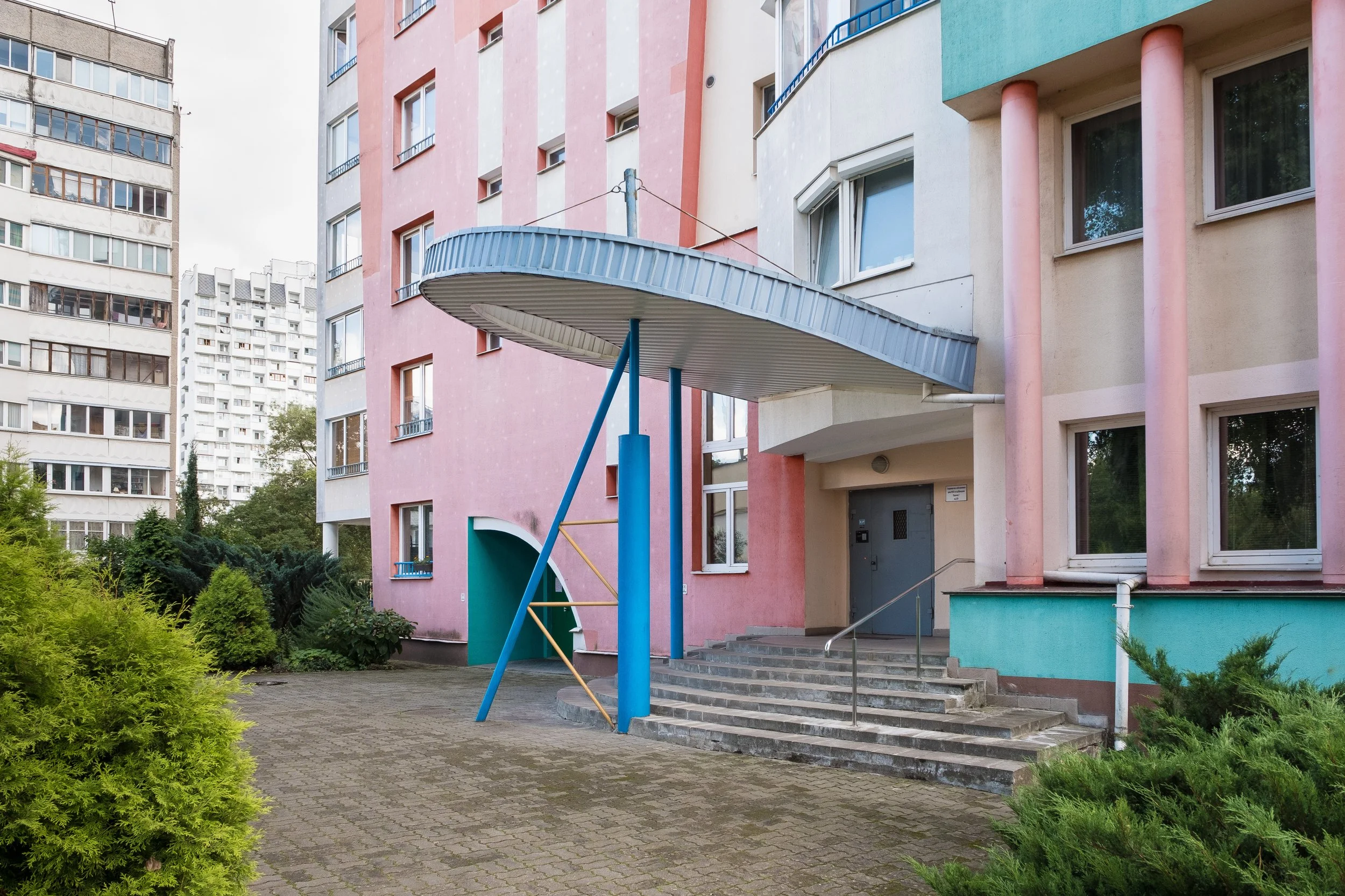 Entrée d'un immeuble résidentiel avec une rampe en métal bleu, un escalier et une entrée principale, entourée de végétation.