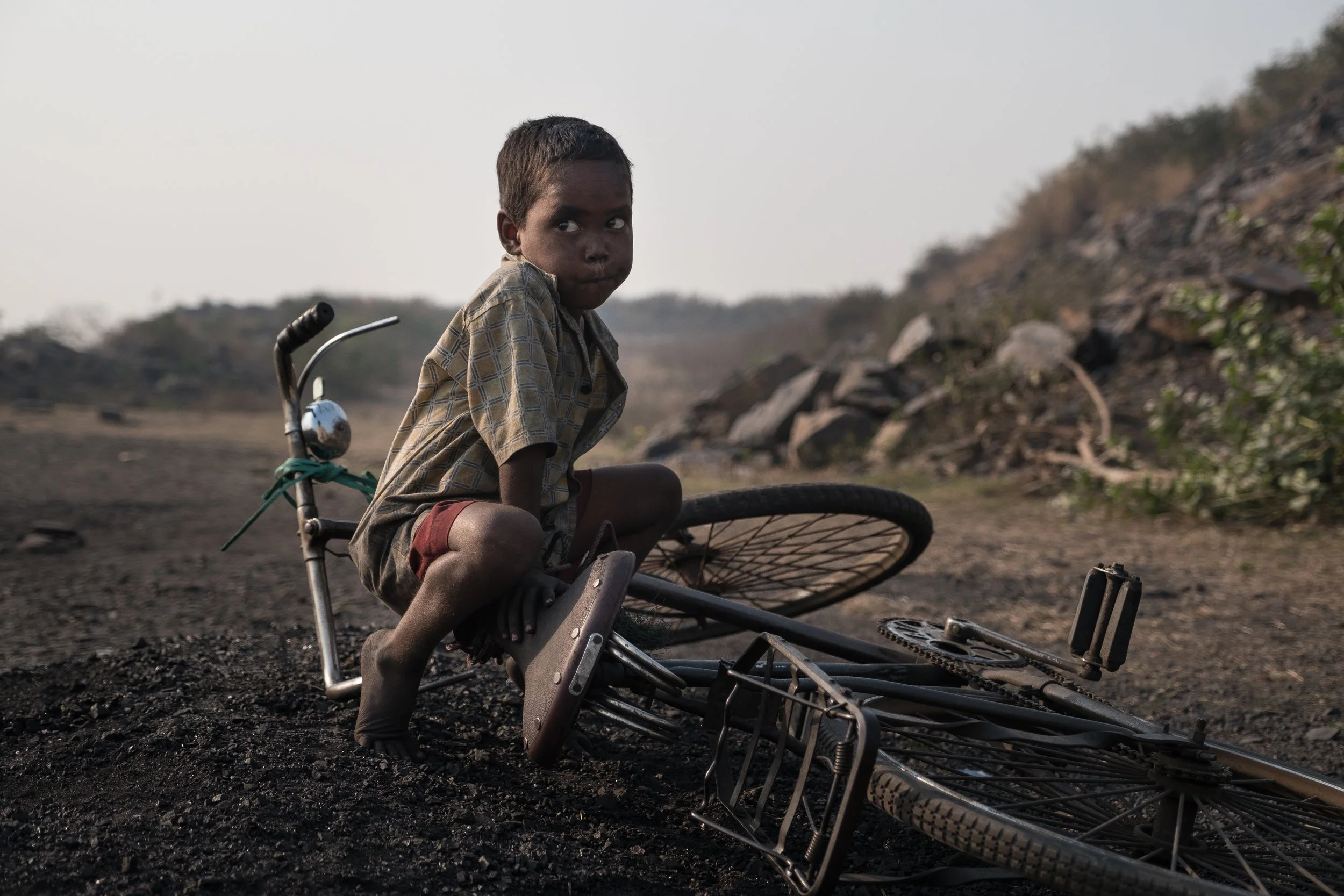 Un jeune garçon à côté d'un vélo tombé sur un sol noir, un paysage rocheux et brouillé en arrière-plan.