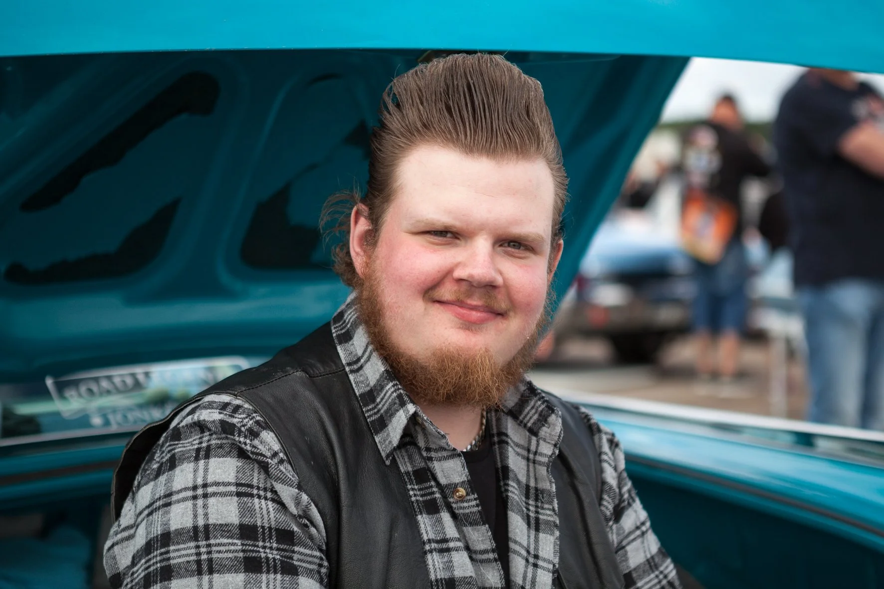 Jeune homme à barbe, cheveux coiffés en arrière, assis sur l'avant d'une voiture ancienne avec le capot levé, portant une chemise à carreaux et un gilet en cuir, lors d'un rassemblement de voitures en Suède.