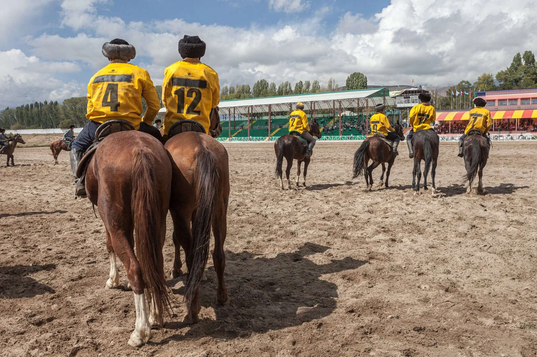 Groupe de cavaliers portant des vestes jaunes avec des numéros, vus de dos, sur un terrain où se dispute un match de kok boru sous un ciel nuageux.