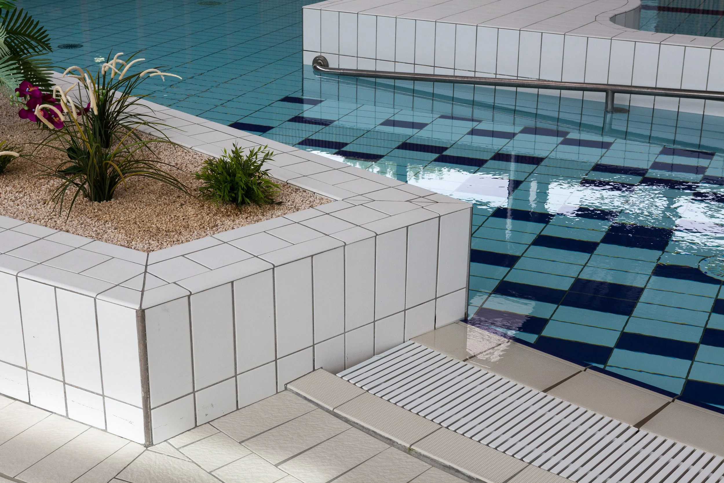 Coin de piscine avec margelle en tuiles blanches, une jardinière en pierres avec des plantes, et le sol en dalles blanches. La piscine a des carreaux bleus et un rampe en métal pour entrer dans l'eau.