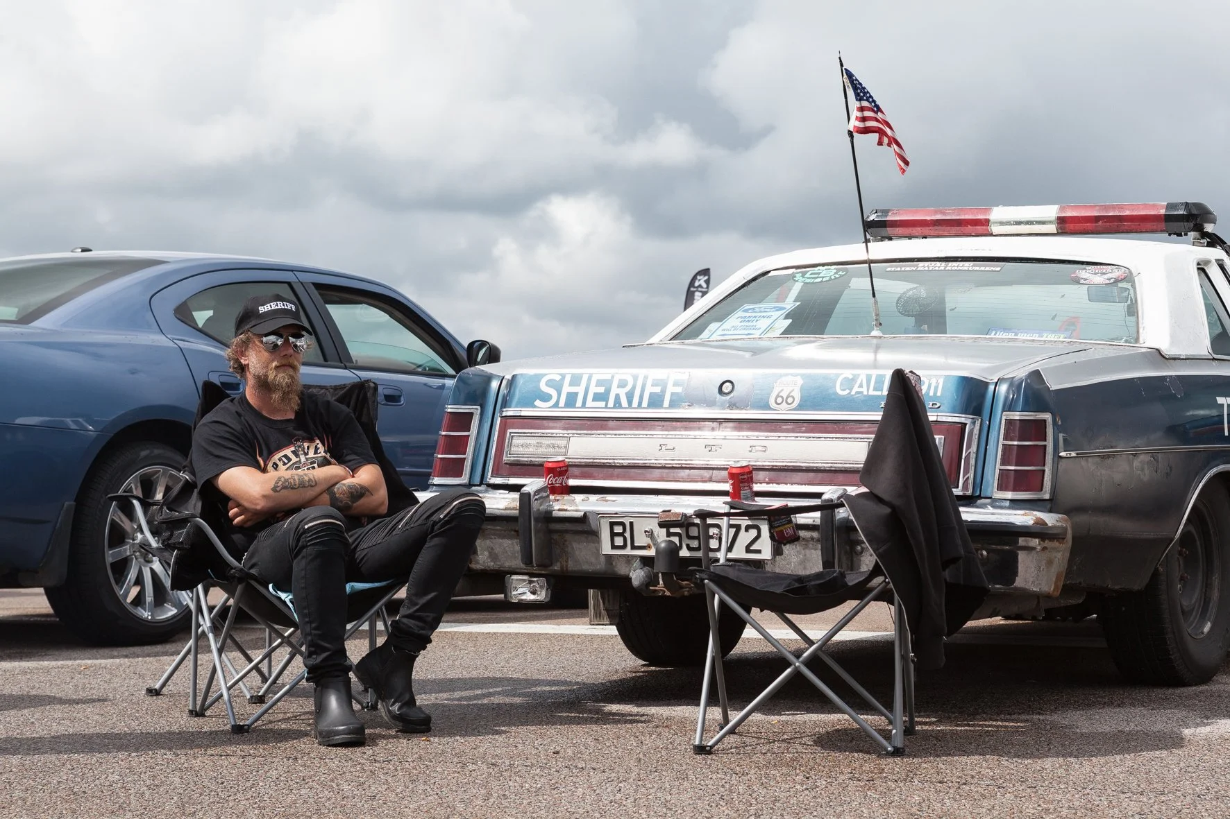 Un homme avec une barbe, lunettes de soleil, casquette et tatouages, assis en chaise pliante devant une voiture de police vintage, aux couleurs de la Californie, avec un drapeau américain planté à l'arrière, dans un parking sous un ciel nuageux.