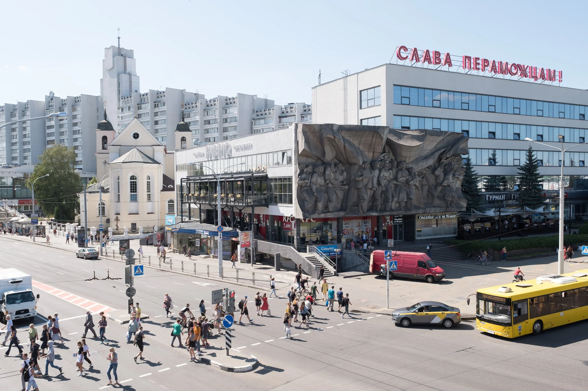 Une rue urbaine avec de nombreux piétons traversant un passage piéton, un bus jaune, un van rouge, des voitures, un immeuble avec une sculpture murale en relief, une petite église blanche avec deux cloches, un bâtiment moderne avec un grand panneau é