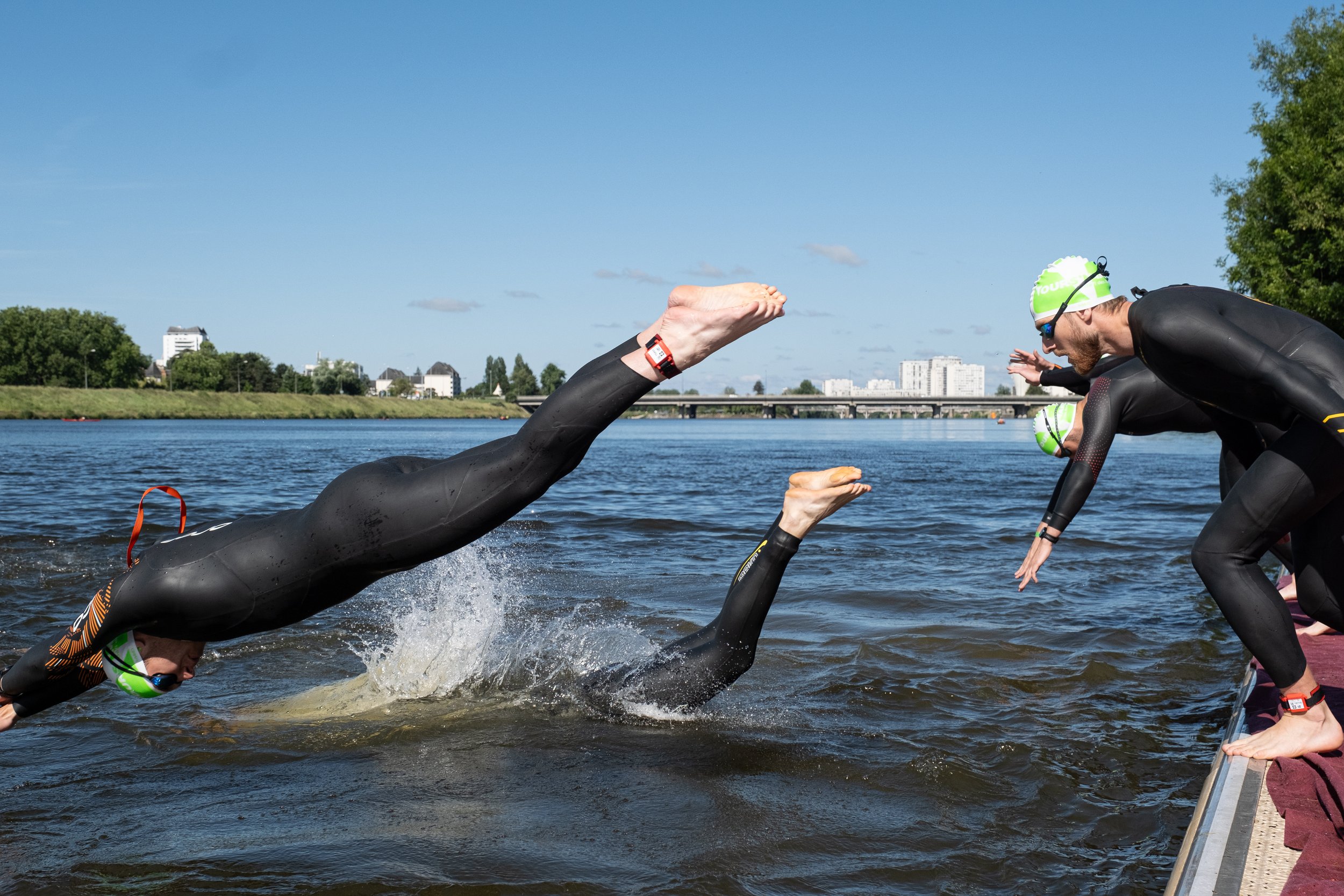 Trois triathlètes en combinaison noire, portant des casques verts, entrant dans l'eau pour une course, l'un d'eux sauté du quai, un autre nageant dans la rivière avec un saut, la scène se déroule par une journée ensoleillée avec un ciel bleu et des b
