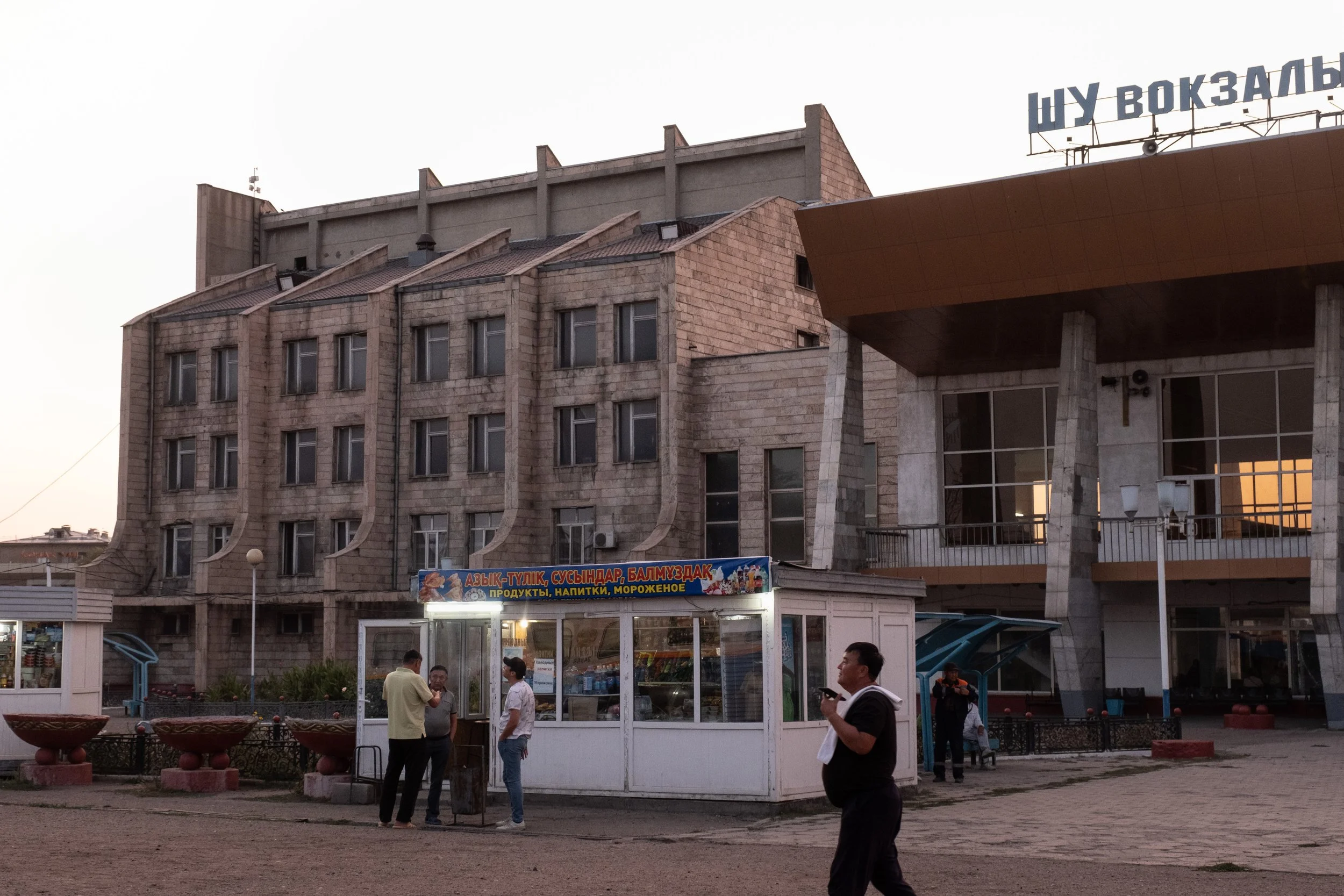 Un groupe de personnes en train de discuter devant une petite boutique de vente de produits, aliments et boissons, située devant un bâtiment brutaliste en pierre avec plusieurs fenêtres, dans une zone urbaine, au coucher du soleil.