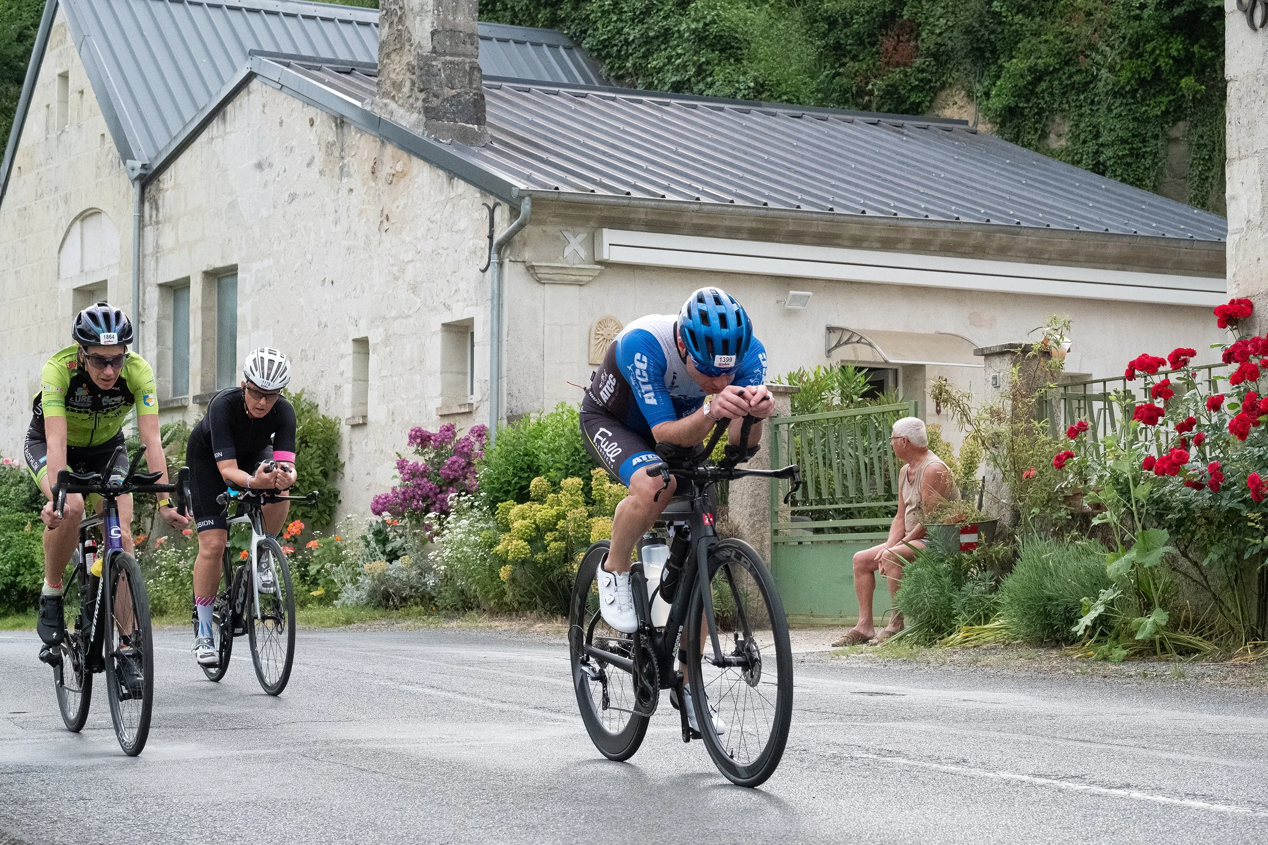 Trois cyclistes en pleine course sur une route de campagne, avec une maison en pierre et un jardin fleuri en arrière-plan, et une personne âgée assise sur un banc près des fleurs.