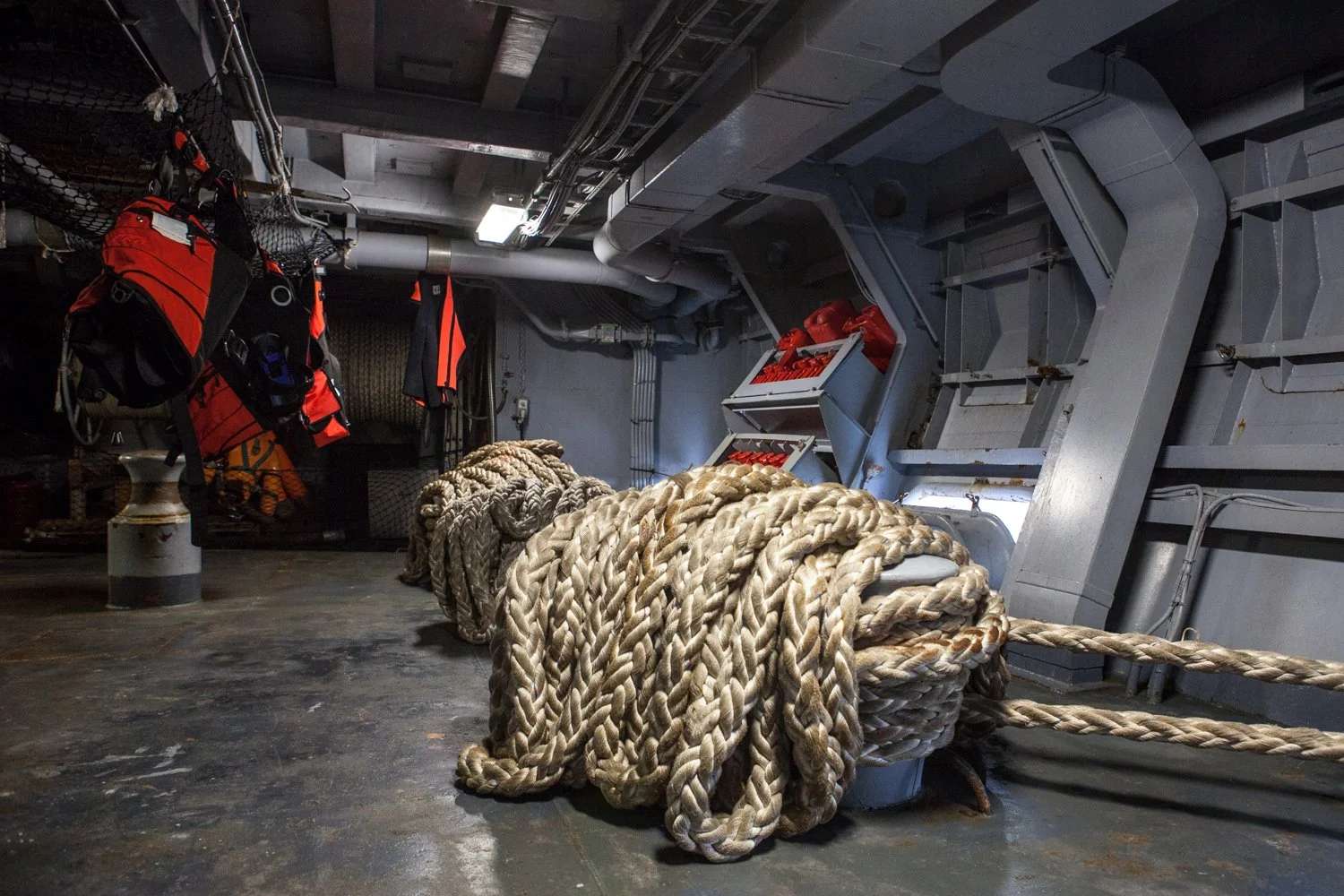 Râteliers avec équipements de sécurité rouge et noir, cordages enroulés en coton épais, dans une salle technique avec murs en métal et tuyaux.