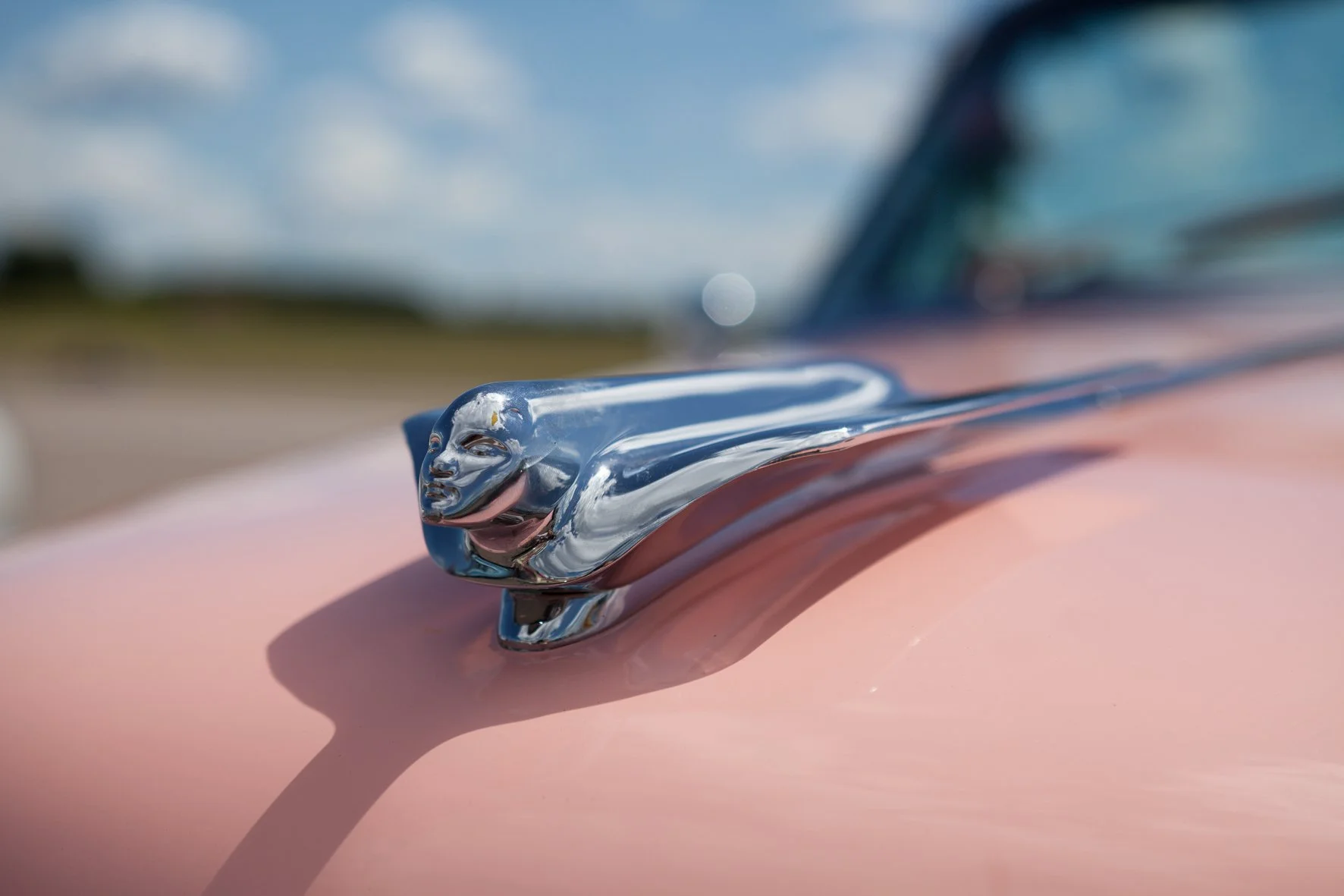 Gros plan d'une mascotte de voiture en forme de rapace en métal chromé sur le capot d'une voiture rose, avec un ciel bleu en arrière-plan.
