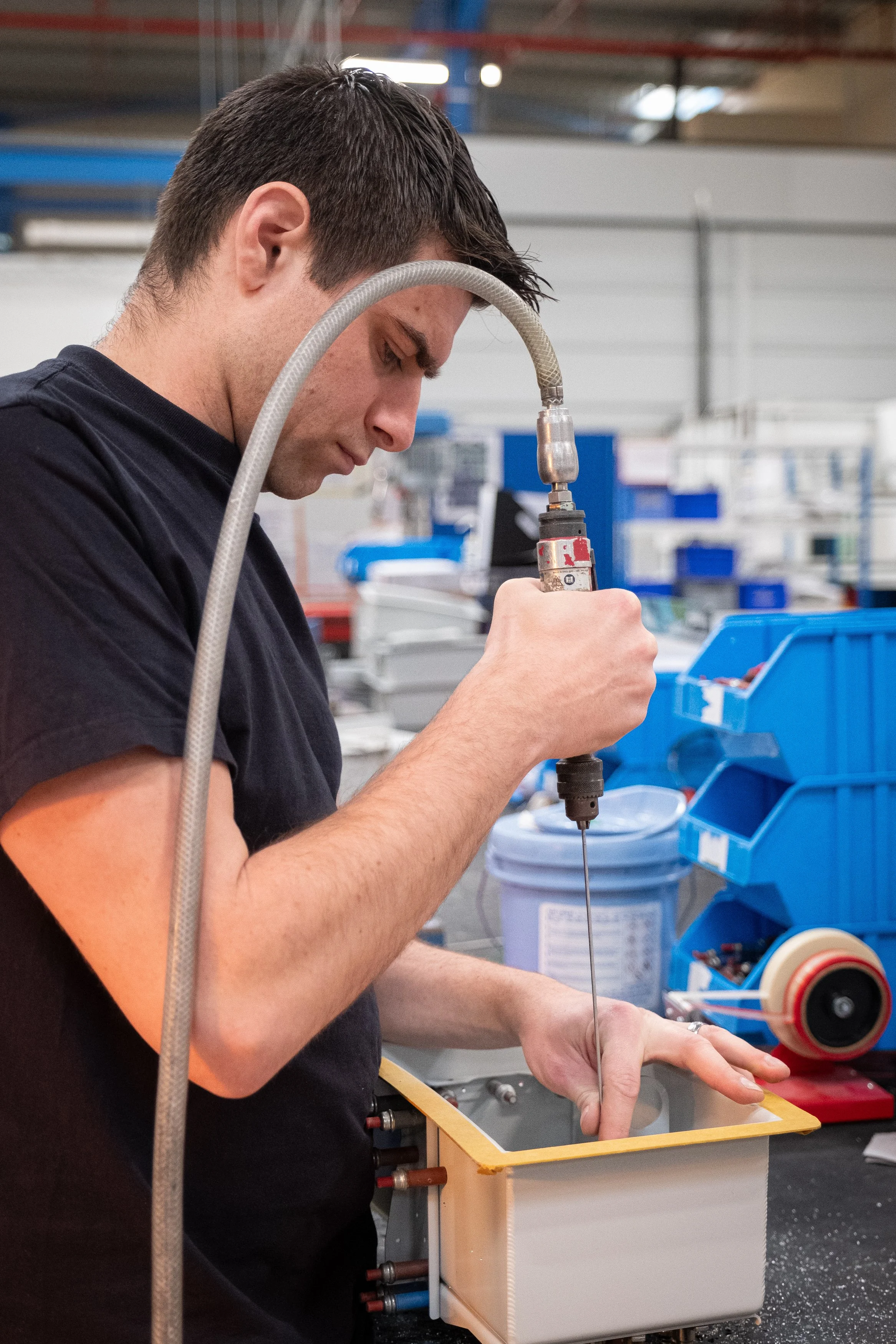 Un homme travaille avec un outil électrique dans un atelier de fabrication. Il manipule un fil ou un câble au-dessus d'une boîte ou un composant technique.