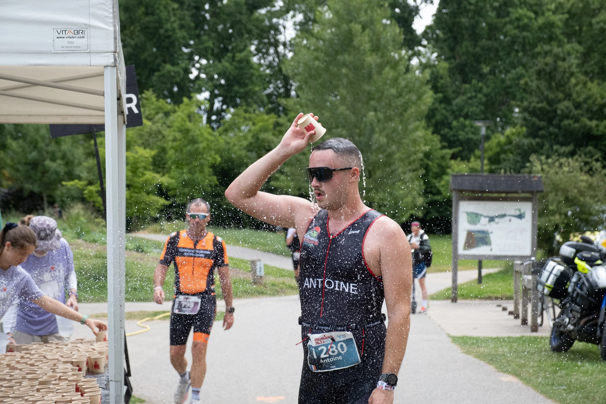 Un homme portant un maillot de course, un casque audio, et des lunettes de soleil, se versant de l'eau sur la tête lors d'une course dans un parc.