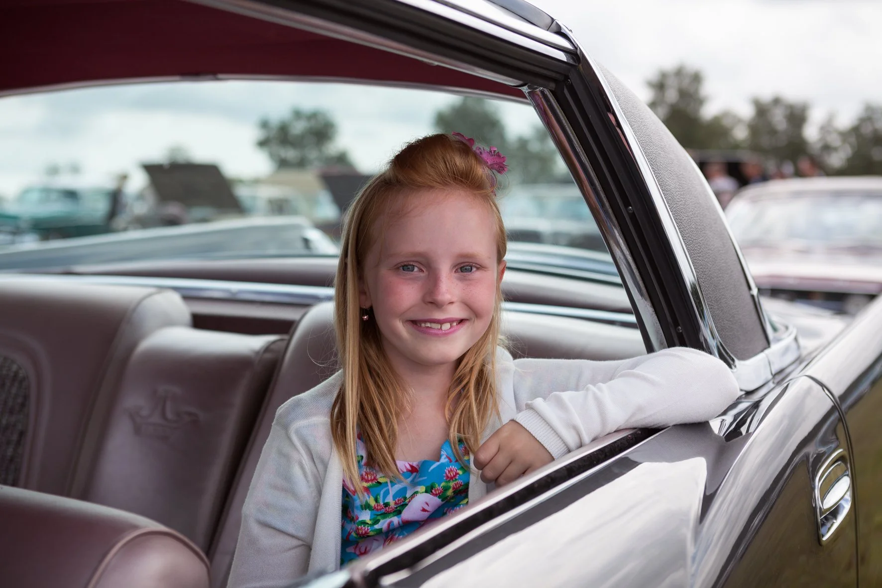Jeune fille rieuse avec cheveux roux et robe colorée, assise dans une voiture classique, lors d'un rassemblement de voitures en Suède.