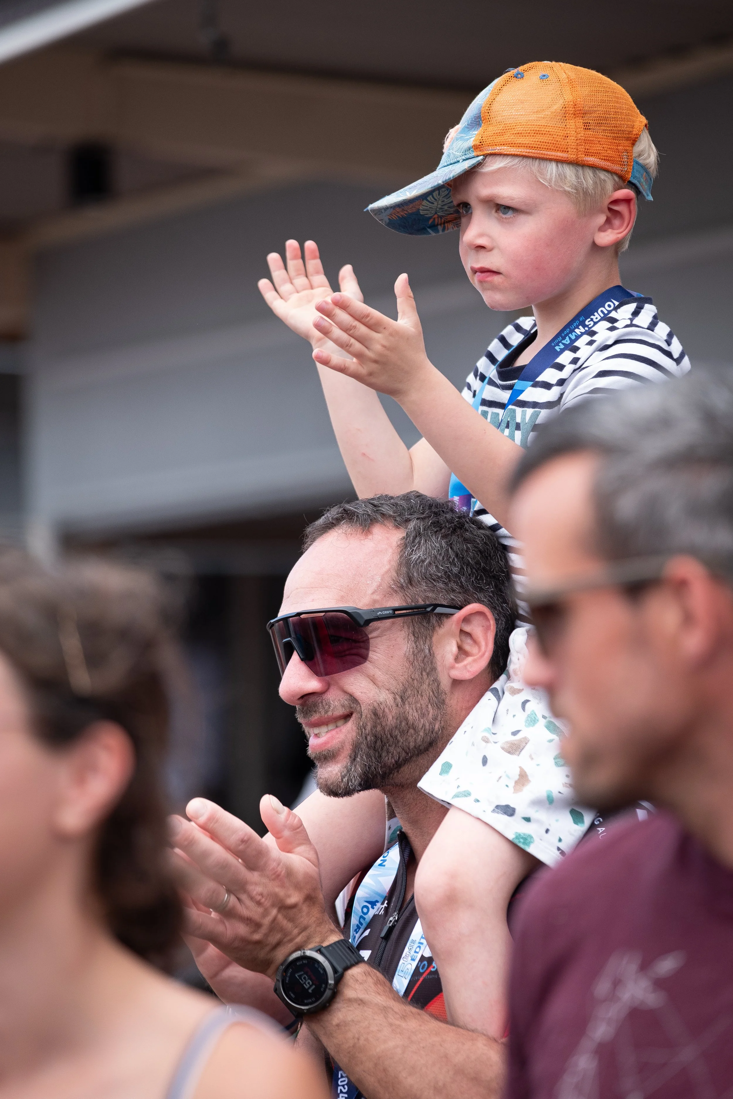Un groupe de personnes regardant vers l'avant, avec un garçon assis sur les épaules d'un homme portant des lunettes de soleil, applaudissant avec d'autres spectateurs dans un environnement extérieur.