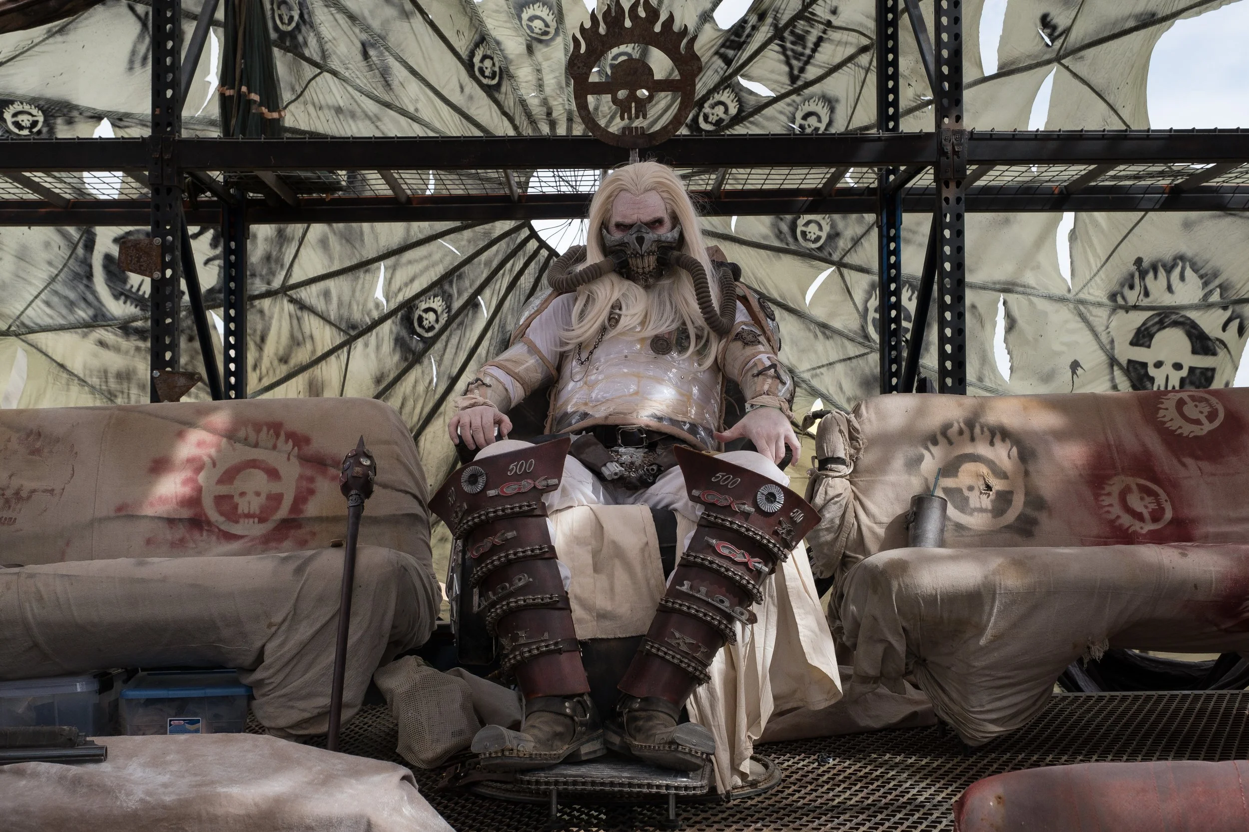 Un personnage en costume de film incarnant Immortal Joe, assis sur un trône, avec un masque facial, des cheveux longs et blancs, dans un décor avec des textiles et des symboles mystérieux à Wasteland.