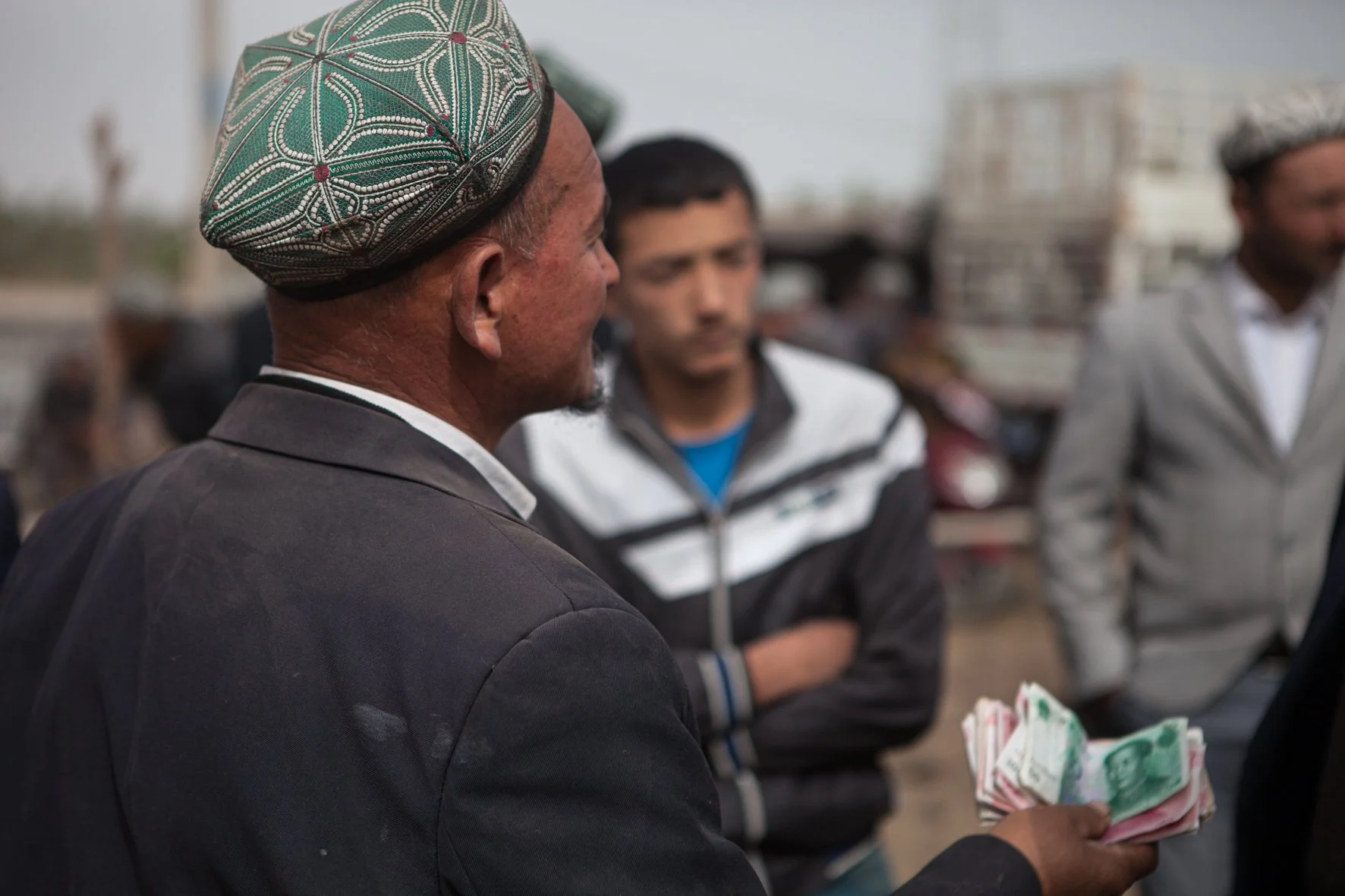 Un homme porte un chapeau traditionnel coloré, tenant des billets de banque, en conversation avec d'autres hommes dans un marché.