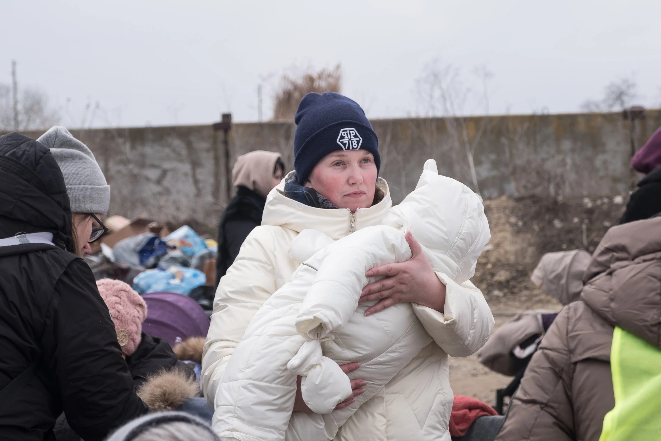 Une femme en manteau blanc tient dans ses bras un bébé enveloppé dans une couverture blanche, dans un contexte de guerre, avec d'autres personnes autour dans un lieu extérieur.