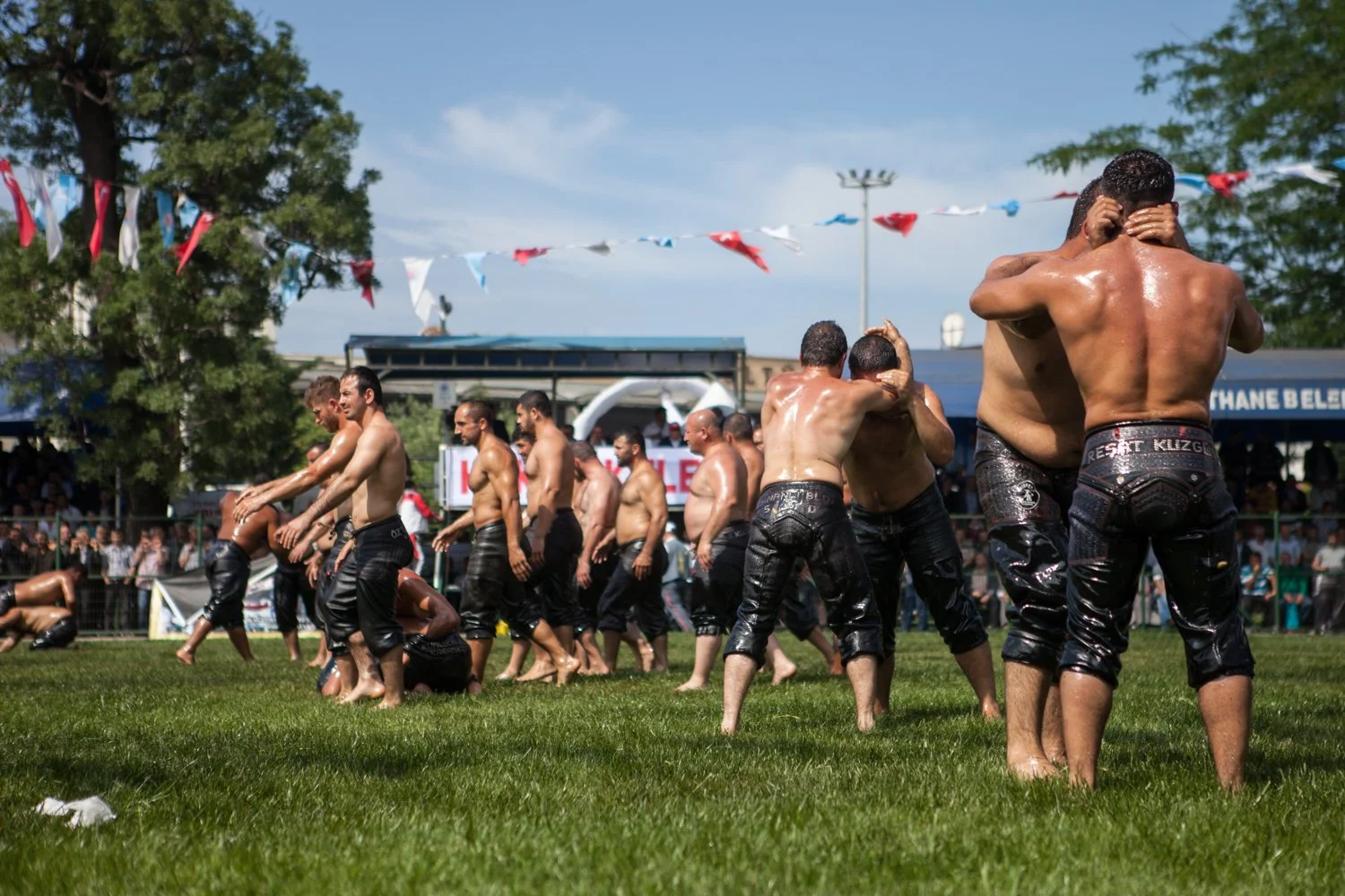 Groupe de personnes en pleine démonstration de lutte traditionnelle turque, vêtues de pantalons en cuir, sur une pelouse lors d'un événement public avec des spectateurs en arrière-plan.