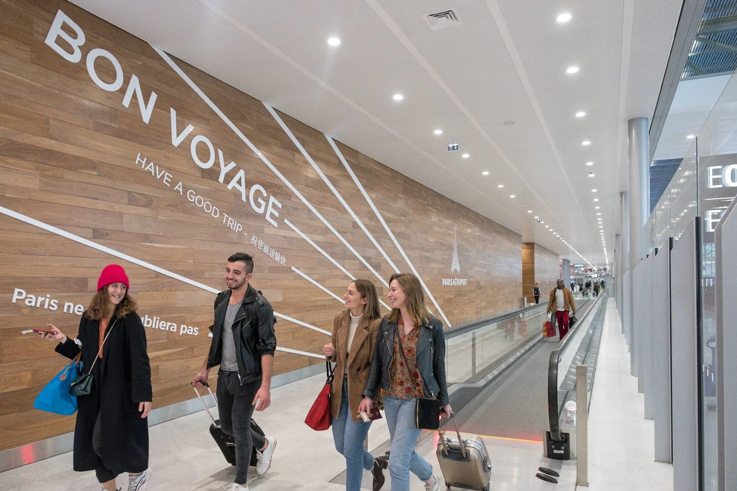 Des passagers souriants marchent dans l'aéroport d'Orly avec un mur en bois décoré d'une citation en français et en anglais, souhaitant un bon voyage.