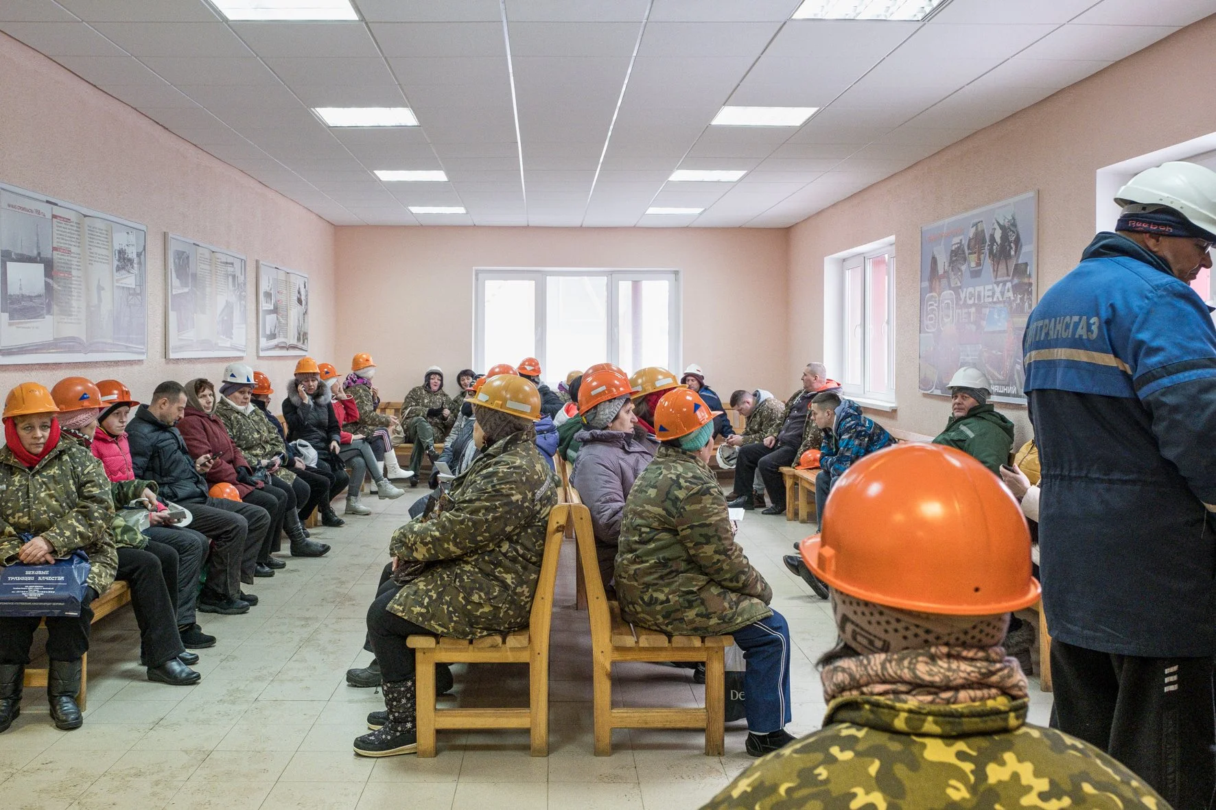 Groupe de personnes portant des casques de sécurité dans une salle d'attente, en manteau à motif camouflage ou en vêtements civils, assises sur des bancs le long des murs, avec des affiches et des fenêtres laissant entrer la lumière.
