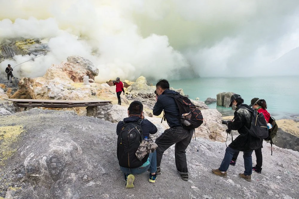 Des personnes prennent des photos et regardent un cratère de volcan en éruption avec de la fumée blanche.