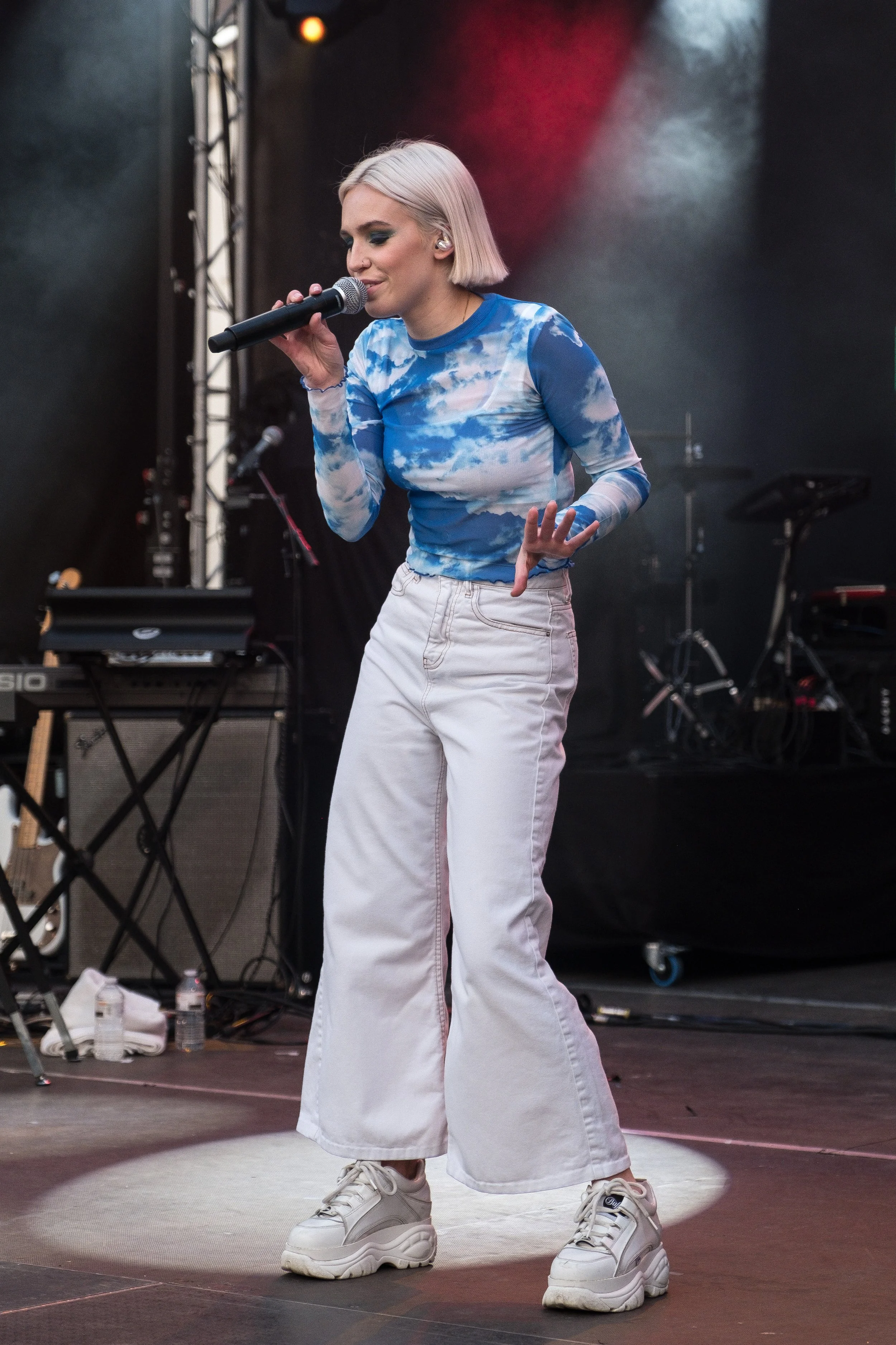 Une jeune femme aux cheveux blonds courts chante en public avec un micro, portant un haut à motif nuages bleus et blancs, un pantalon blanc large et des chaussures blanches, sur une scène de concert.