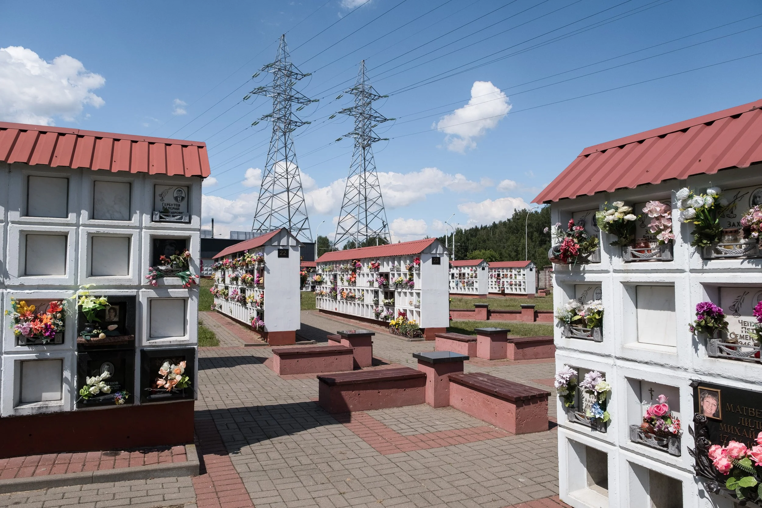 Cimetière avec des mausolées blancs ornés de fleurs artificielles, des allées pavées, et des pylônes électriques en arrière-plan sous un ciel bleu avec quelques nuages à Minsk.