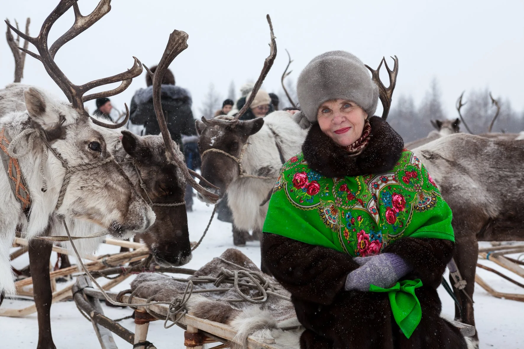 Une femme vêtue de vêtements traditionnels russes avec un chapeau en fourrure, entourée de rennes dans un paysage hivernal enneigé.