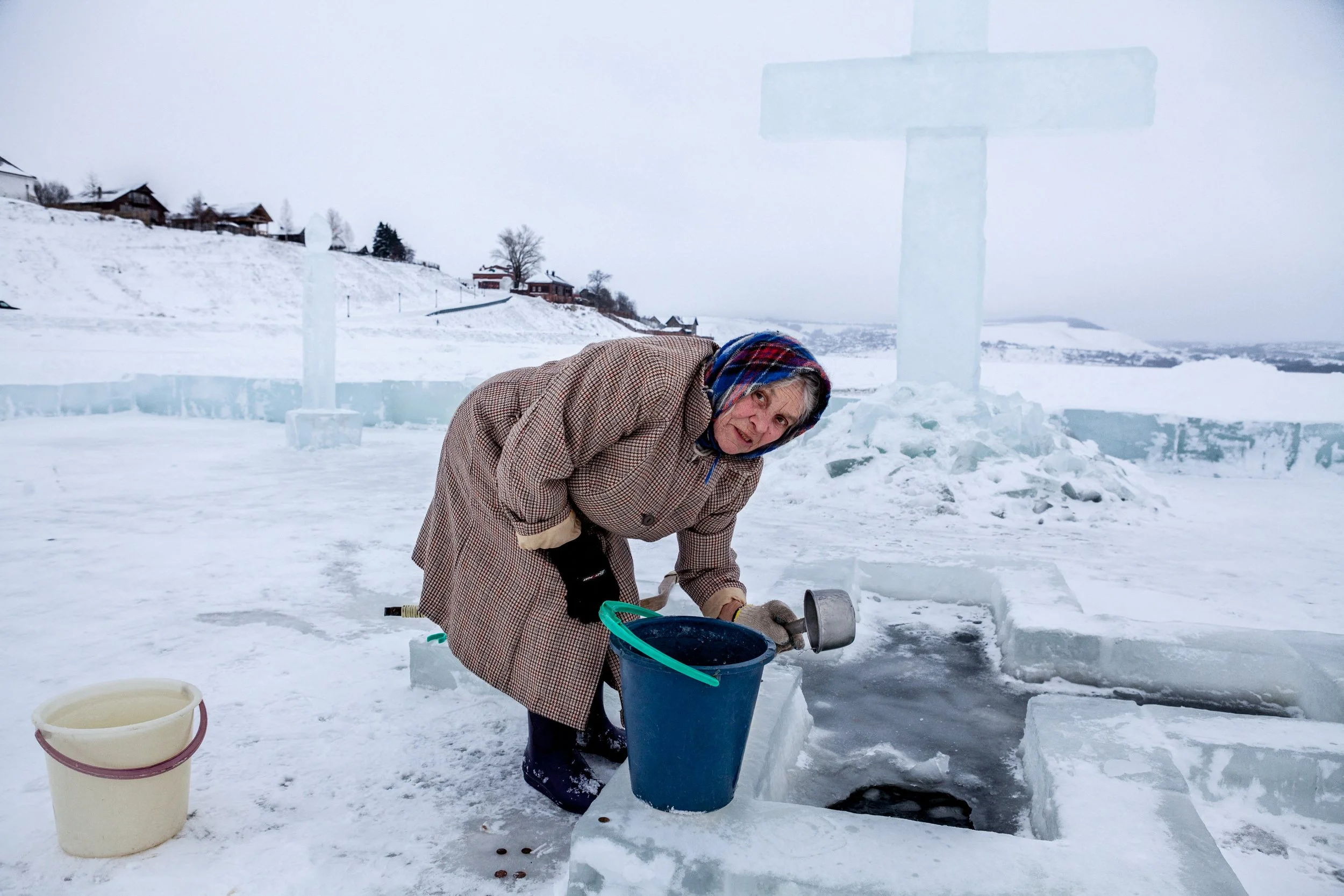 Une femme âgée en vêtements d'hiver, avec un foulard, rempli un seau avec un outil pour puiser de l'eau dans un trou creusé dans la glace en plein hiver, sur un paysage enneigé avec des maisons en arrière-plan, en Russie.