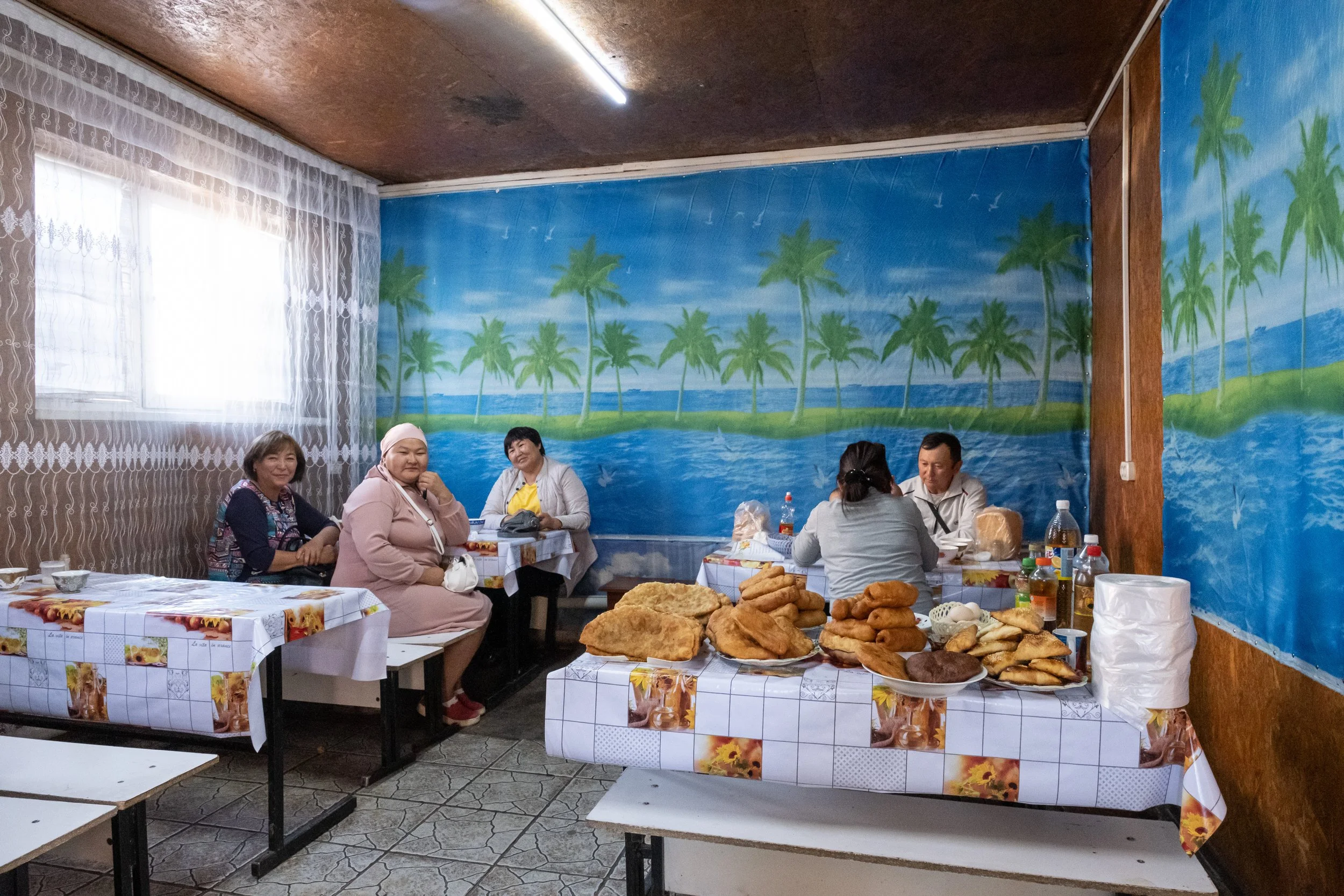 Groupe de personnes assises autour de tables dans une salle décorée d'un mur peint avec des palmiers et un paysage tropical. Tables avec des plats de nourriture, notamment des pains et autres aliments, ainsi que des boissons au Kazakhstan.