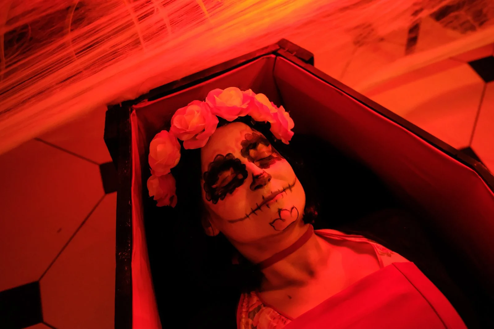 Une femme avec un maquillage de style calavera, portant une couronne de roses, allongée dans un cercueil. L'ambiance est éclairée en rouge pour un effet dramatique ou festif. Le décor évoque un Día de los Muertos.
