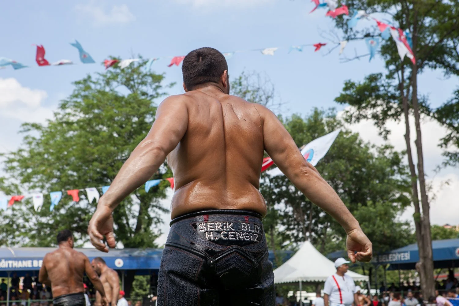 Un homme torse nu avec des bras puissants regarde sur le côté lors d'une compétition de lutte en plein air, avec des fanions colorés en arrière-plan.