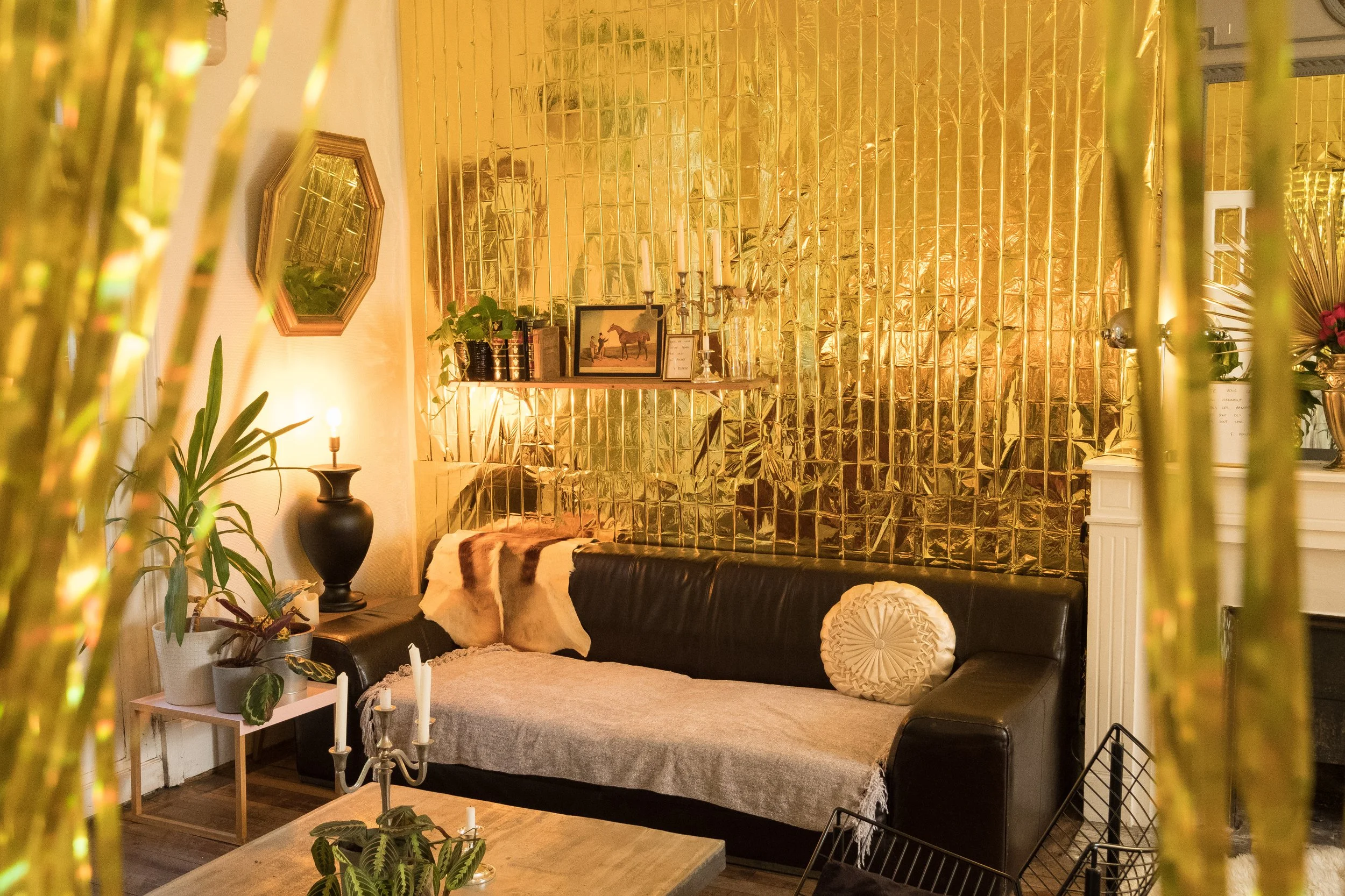 Salon avec canapé noir, tapis beige, mur en miroir doré, bibelots, plantes, et chandelier blanc.