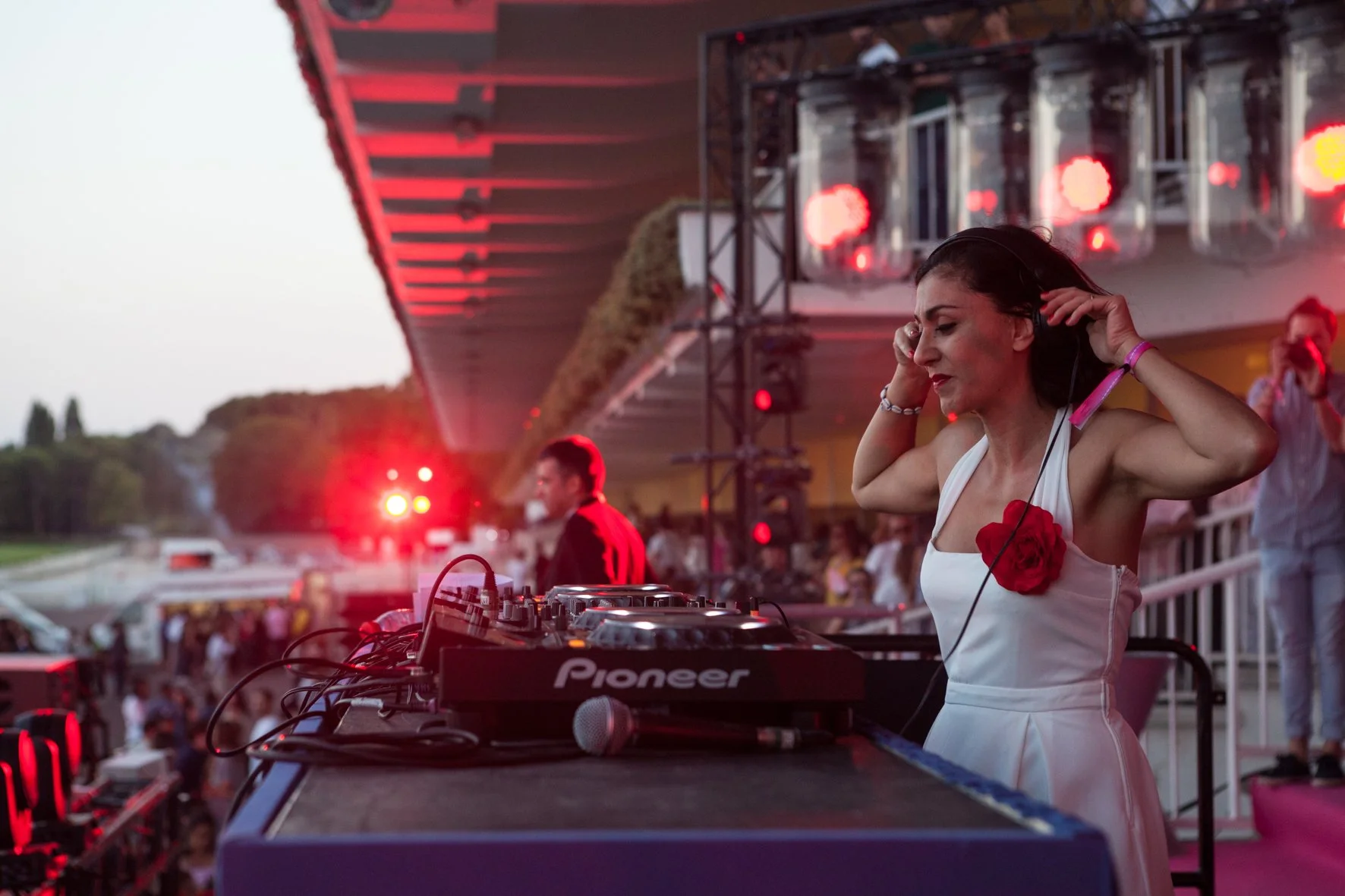 Une DJ femme en robe blanche avec une rose rouge à la poitrine, portant un casque et jouant de la musique lors d'un événement au coucher du soleil, avec une foule en arrière-plan.