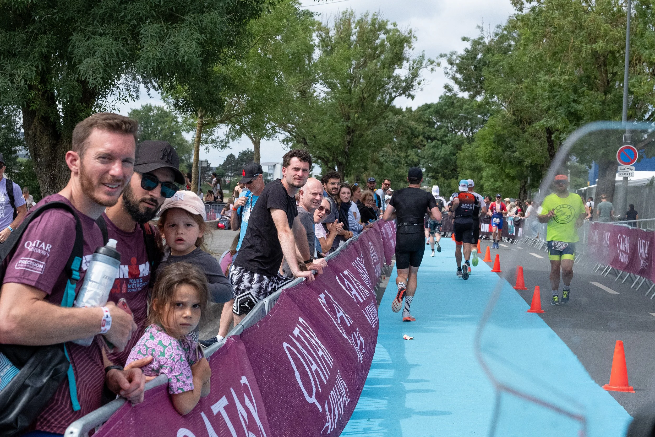 Spectateurs regardant un triathlon sur une piste bleue, avec des arbres en fond et des barrières violettes.