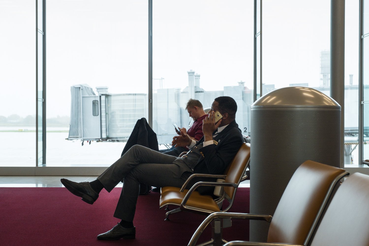 Deux hommes assis dans une salle d'attente d'aéroport, l'un utilisant un téléphone portable et l'autre regardant son téléphone.