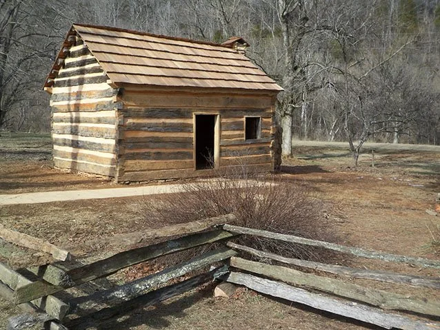 Old log cabin with a shingled roof in a wooded area, surrounded by a weathered wooden fence and leafless trees.