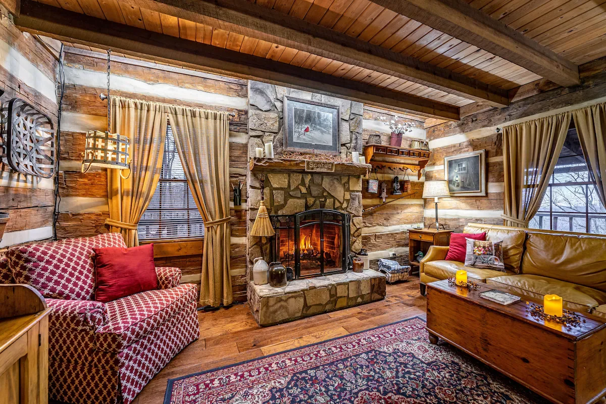 Cozy rustic living room with wood-paneled walls, a stone fireplace with a fire burning, a red patterned armchair with a red pillow, a yellow leather sofa with decorative pillows, a wooden coffee table with candles, and large windows with gold curtains, creating a warm inviting atmosphere.